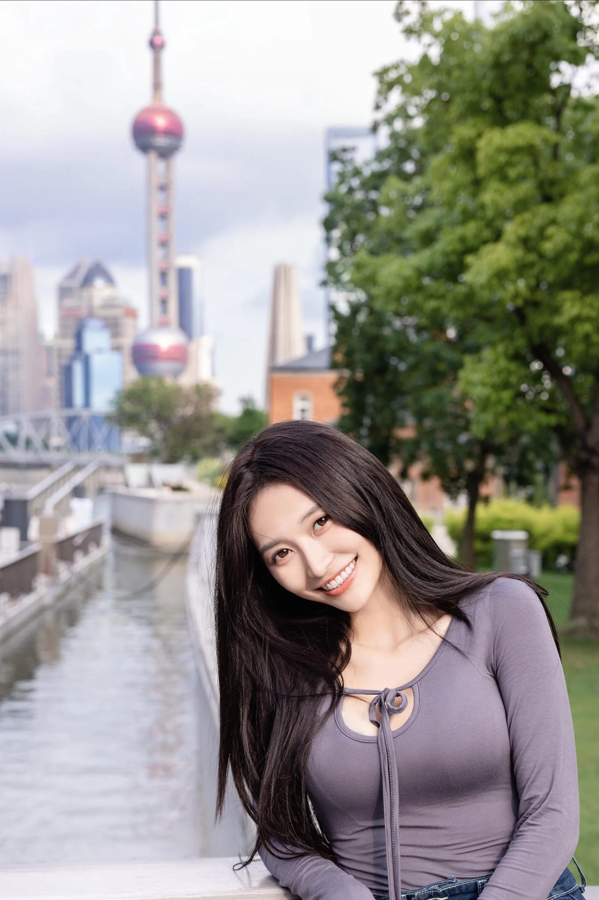 A smiling woman with long dark hair poses outdoors near a canal with a city skyline and the Oriental Pearl Tower in the background.