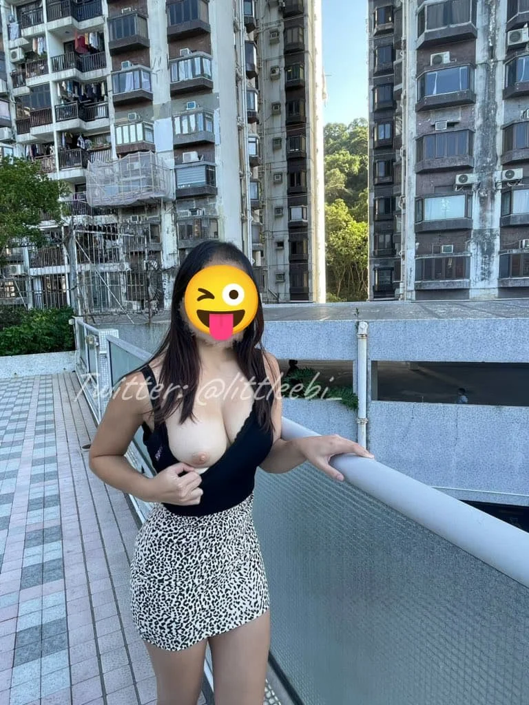 A woman standing on a balcony of an apartment building, wearing a black top and a leopard print skirt, with her face covered by a winking sticker emoji.
