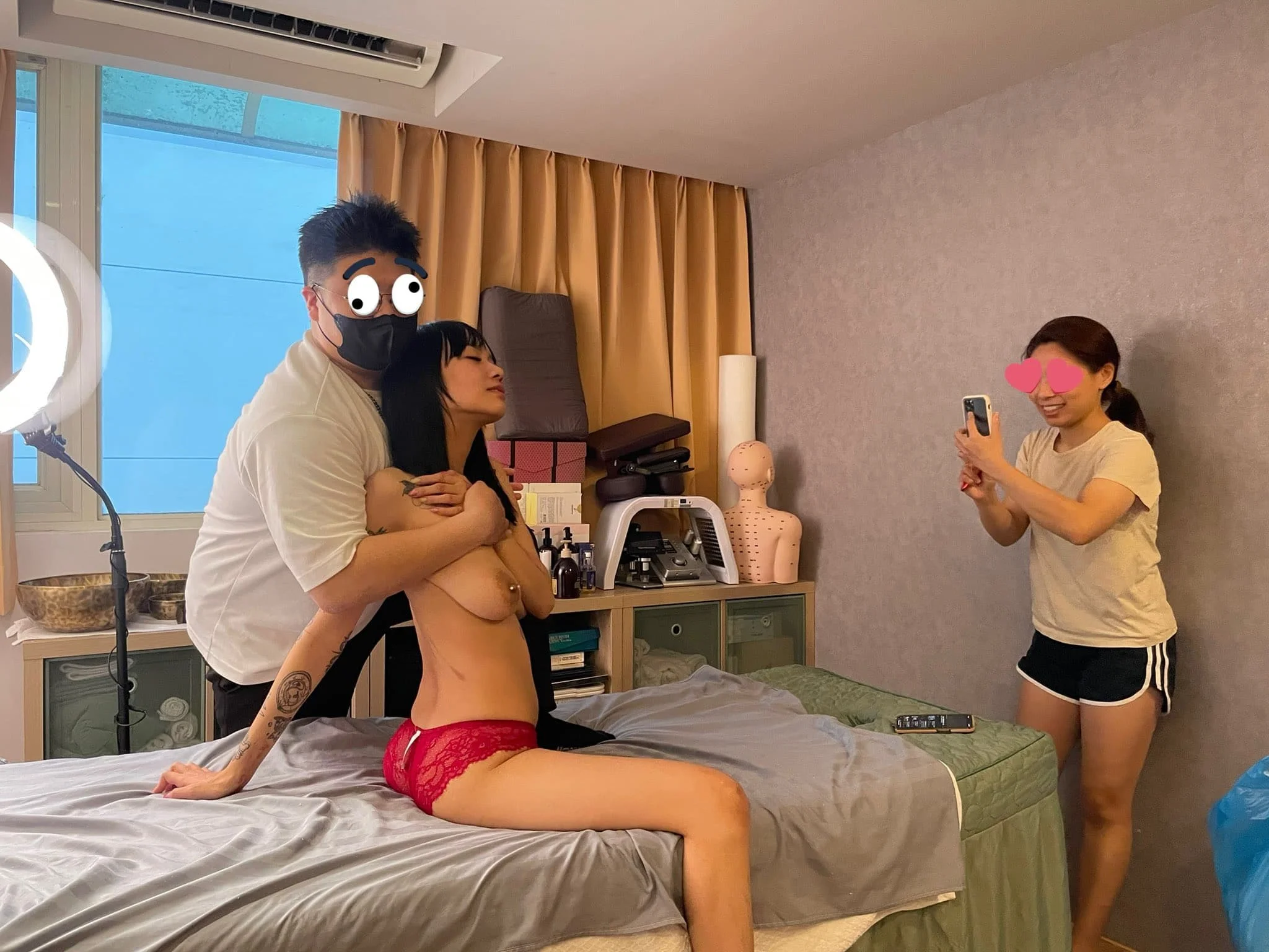 A woman in red lingerie is posing on a bed while a man with cartoon eyes and a mask stands behind her with arms around her shoulders. Another woman in casual clothes is taking a photo with her phone. The setting appears to be a bedroom with medical t