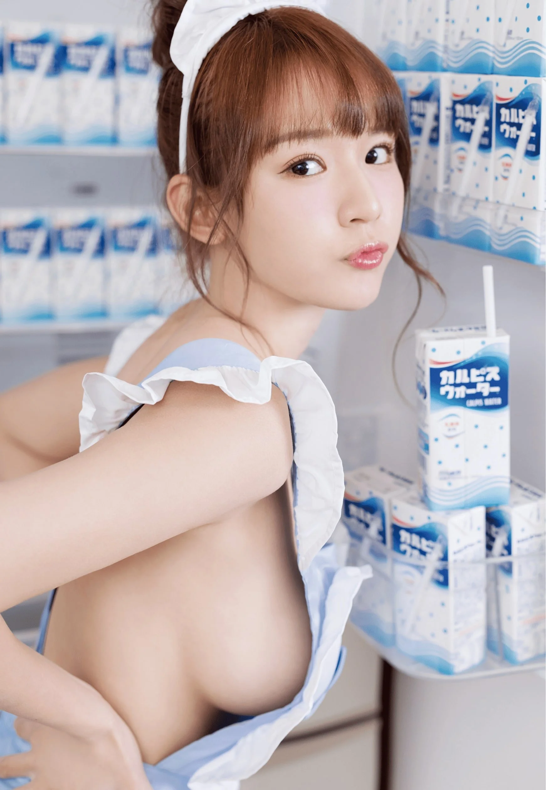 A woman with brown hair tied up, wearing a light blue dress with ruffles, poses next to a display of Japanese beverage cartons. The cartons are labeled with "Calpis Water" in blue text and feature a straw.