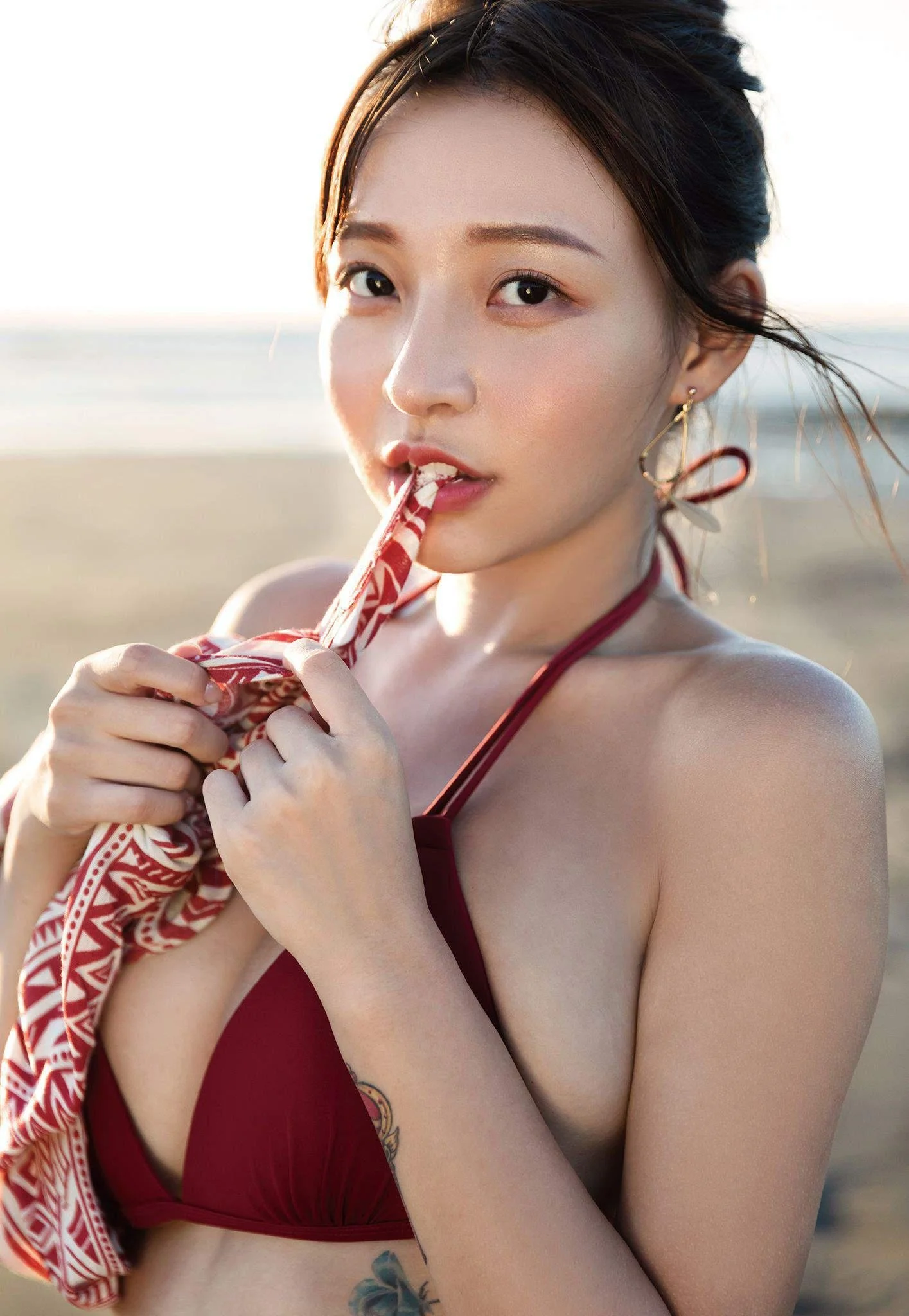A woman in a red bikini is on the beach, holding a patterned fabric and biting it while looking at the camera with her hair blowing in the wind.