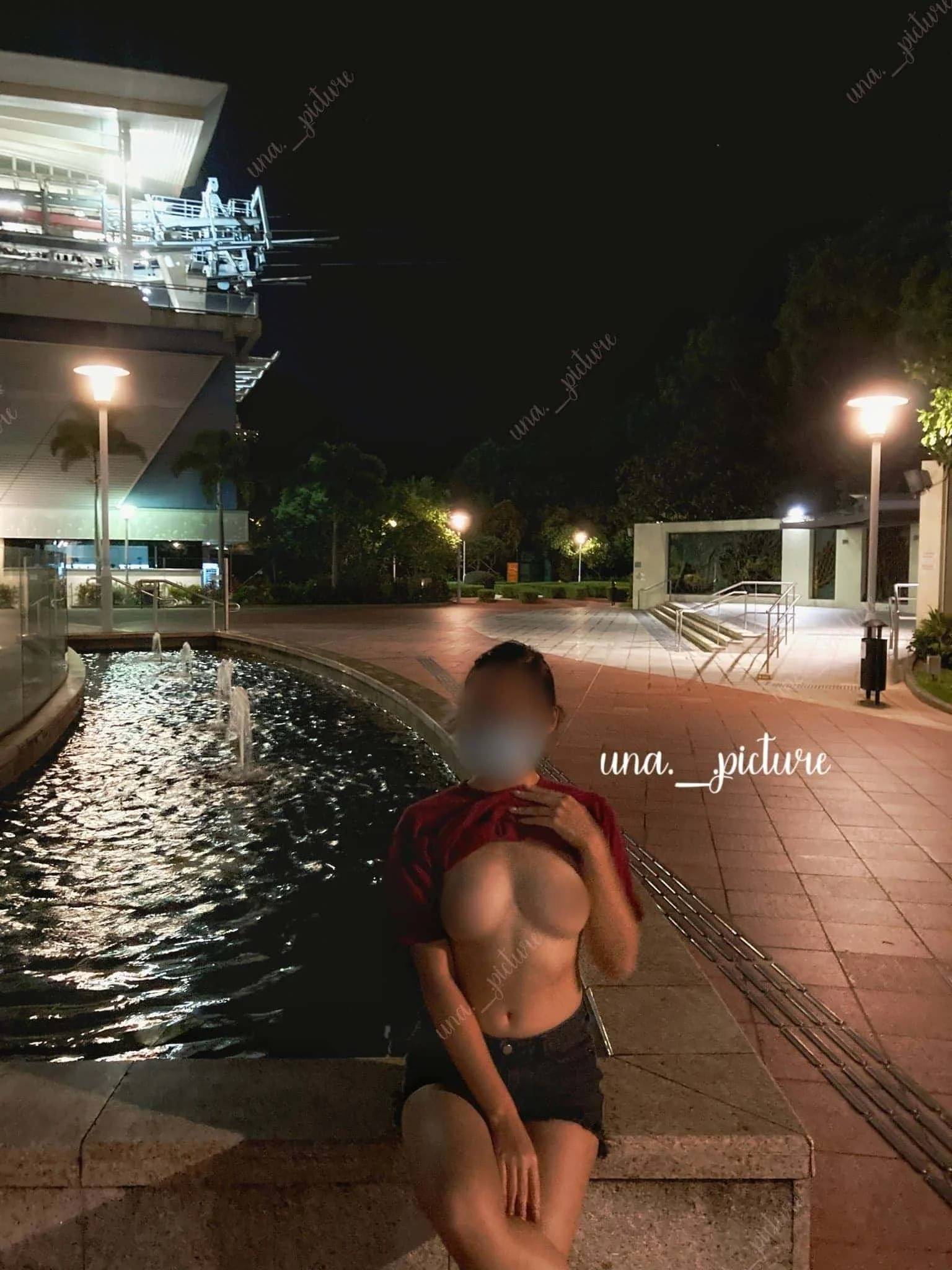 A woman with blurred face and exposed chest sitting outdoors at night near a water feature and lit street lamps.