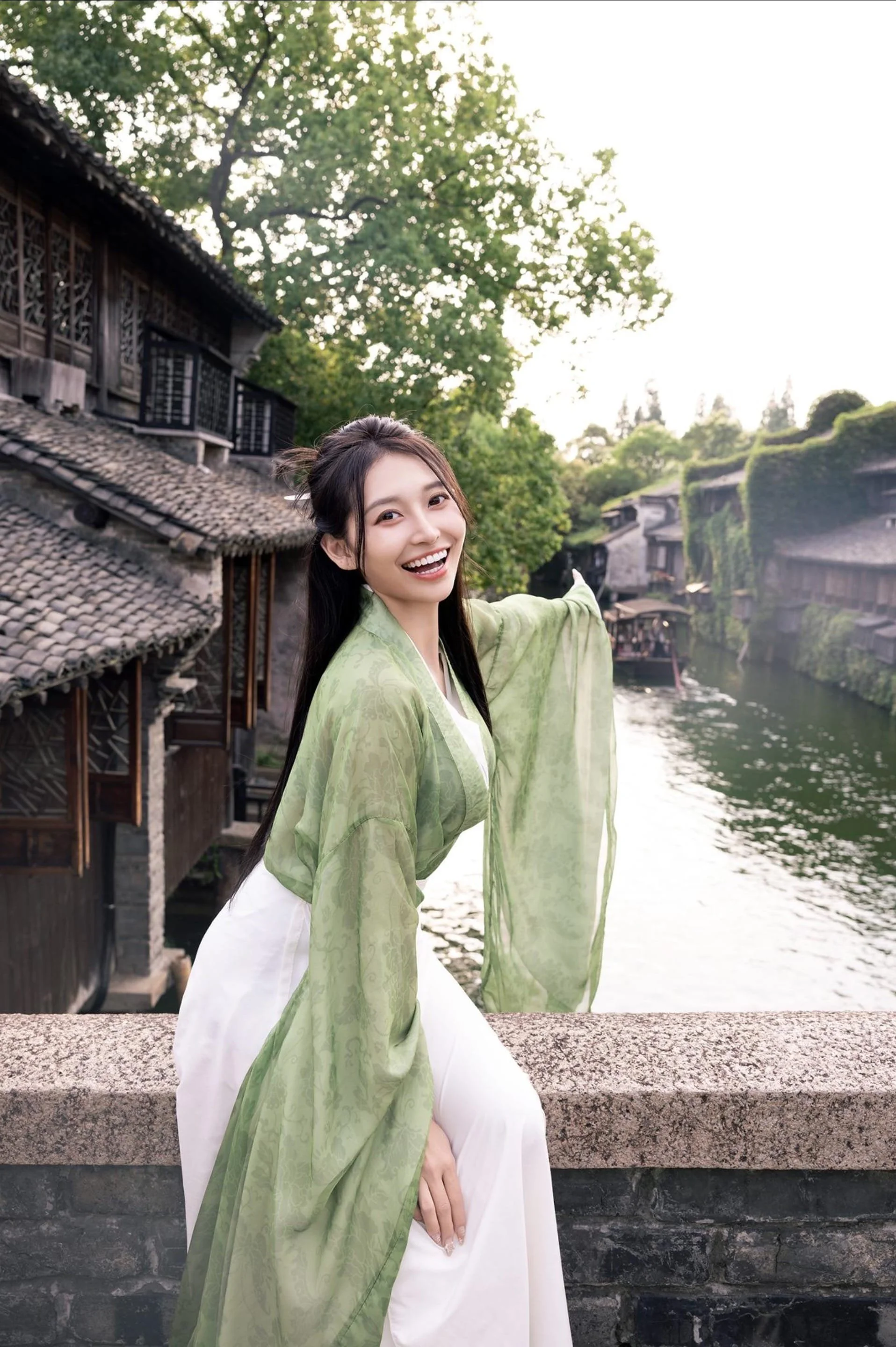 A woman in traditional Chinese attire with a green robe and white skirt, smiling and pointing towards a waterway with historic Chinese-style buildings on both sides.