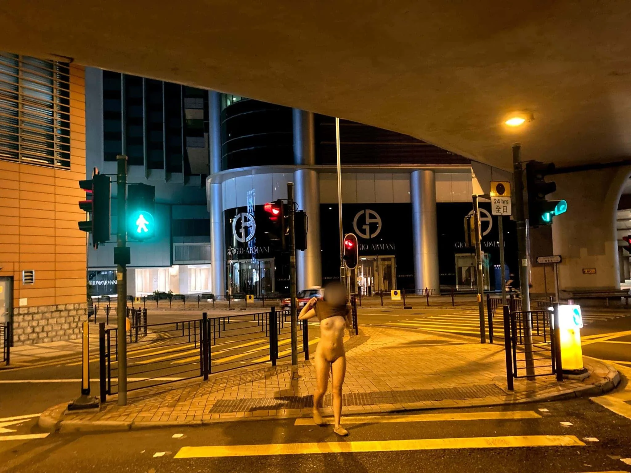 Naked man standing on a crosswalk at night with traffic lights and a Giorgio Armani store in the background.