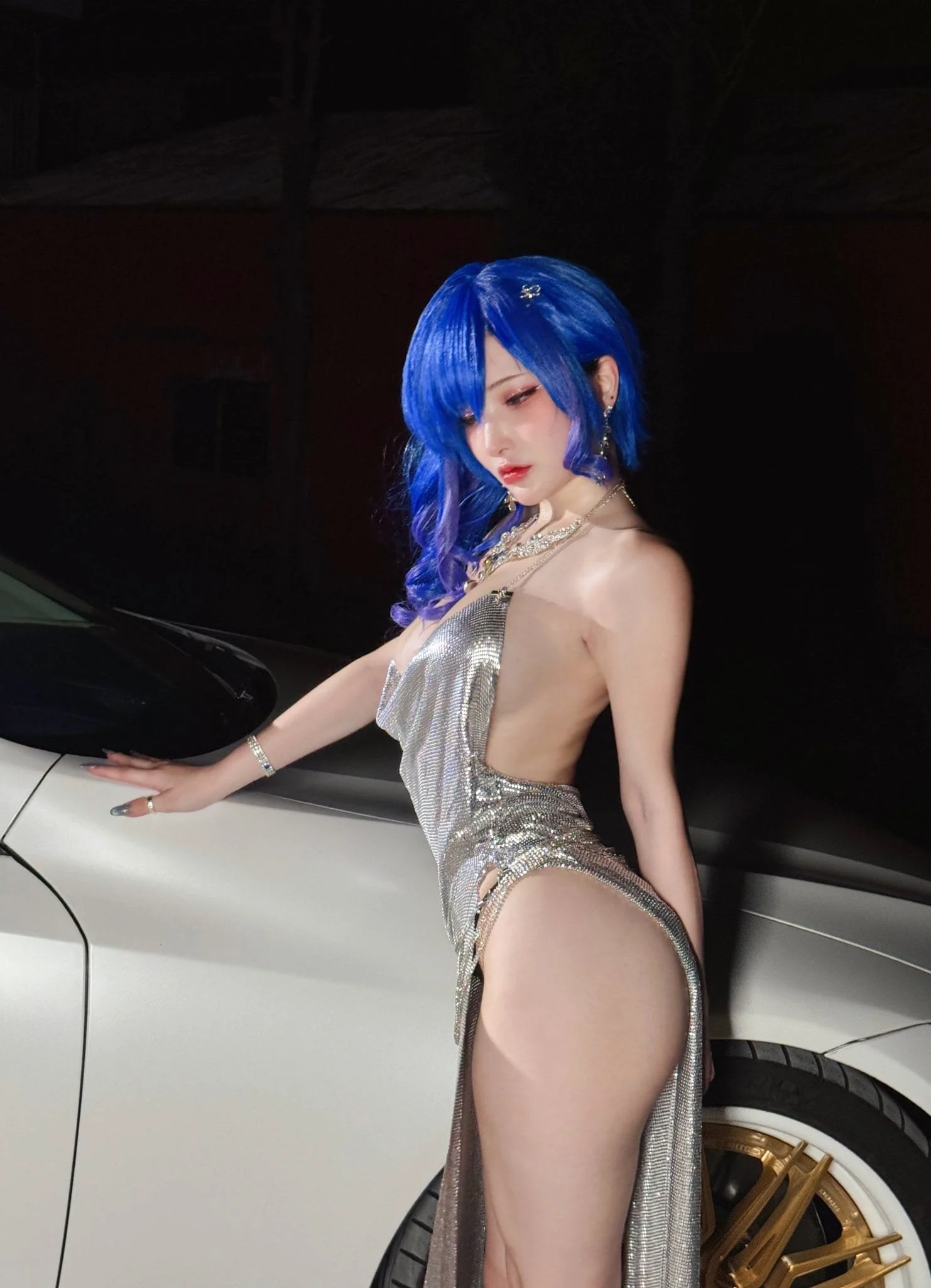 A woman with bright blue hair and silver earrings, wearing a revealing, shiny silver dress, leaning on a white luxury car with gold rims, in a nighttime setting.