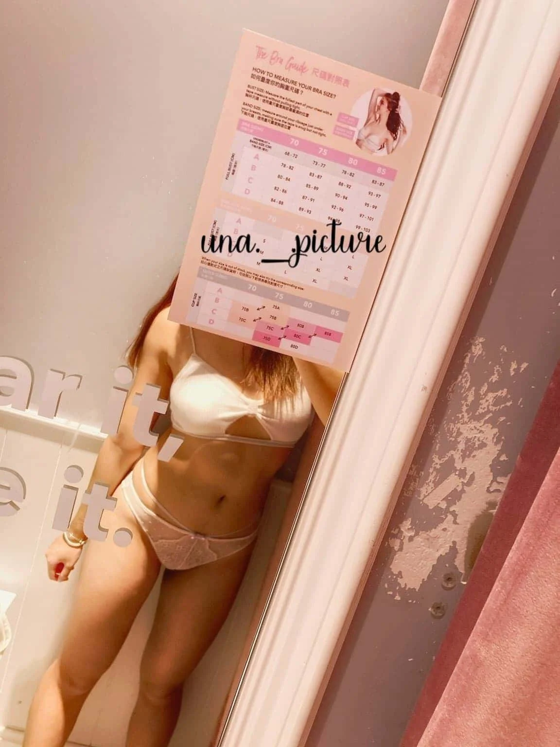 A woman wearing white lingerie is taking a mirror selfie in a fitting room, holding a pink chart that covers her face. The wall behind her is gray with peeling paint, and the mirror has a partial white decorative border.