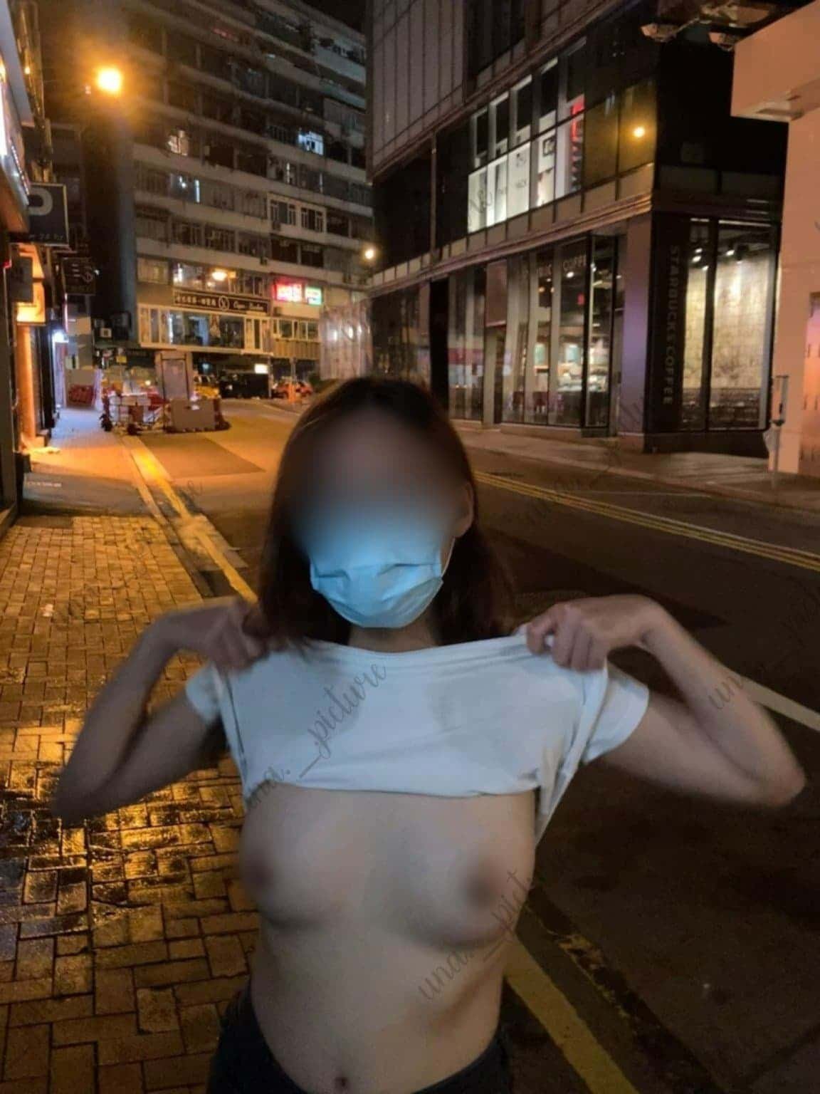A woman wearing a face mask stands on a city street at night, lifting her shirt to expose her upper body. She is in front of buildings with illuminated windows and streetlights.