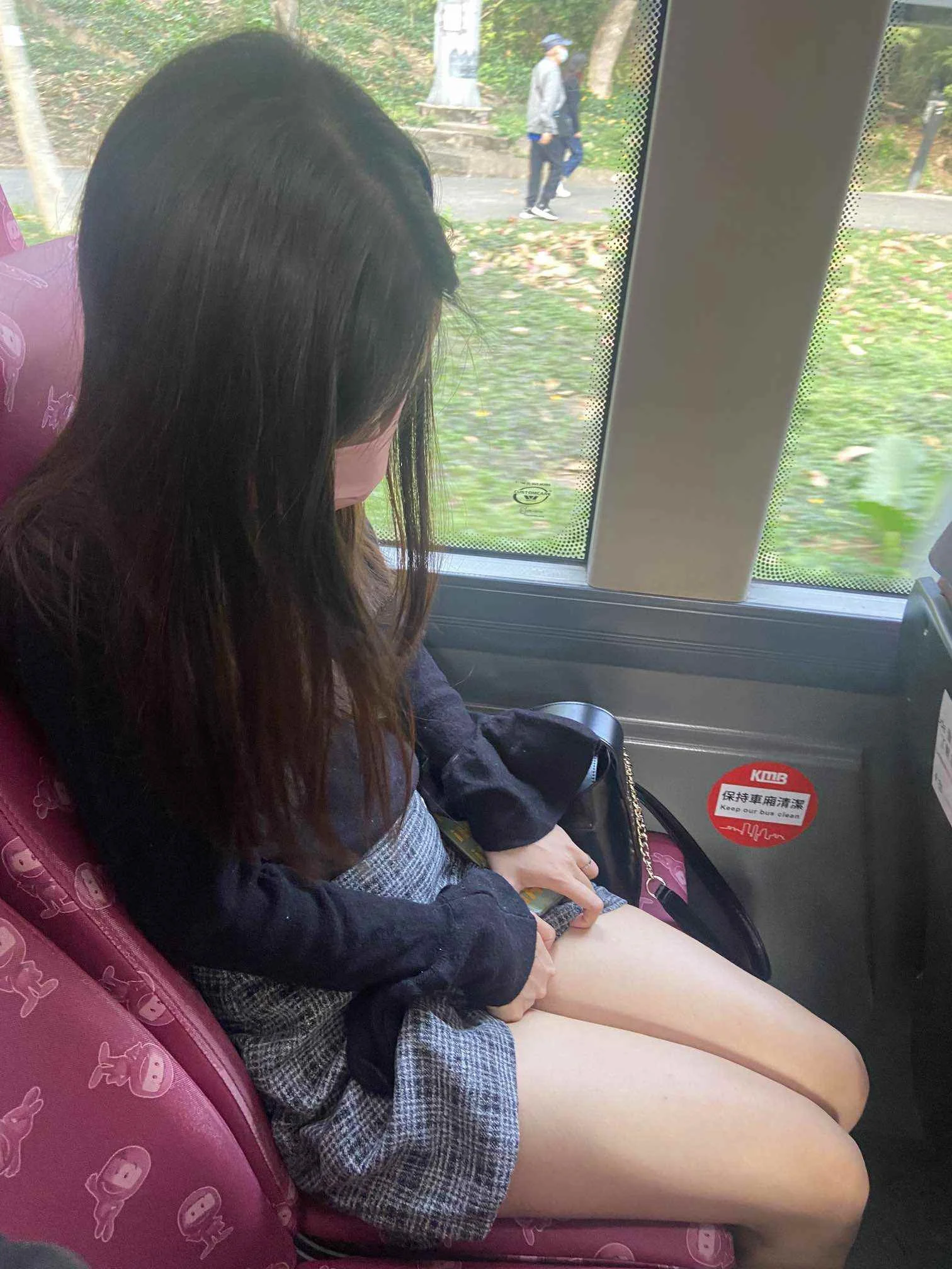 A young woman with long dark hair is sitting on a pink seat with cartoon character designs on a bus, looking down at her phone. Outside the window, two people are walking in a park with fallen leaves and trees.