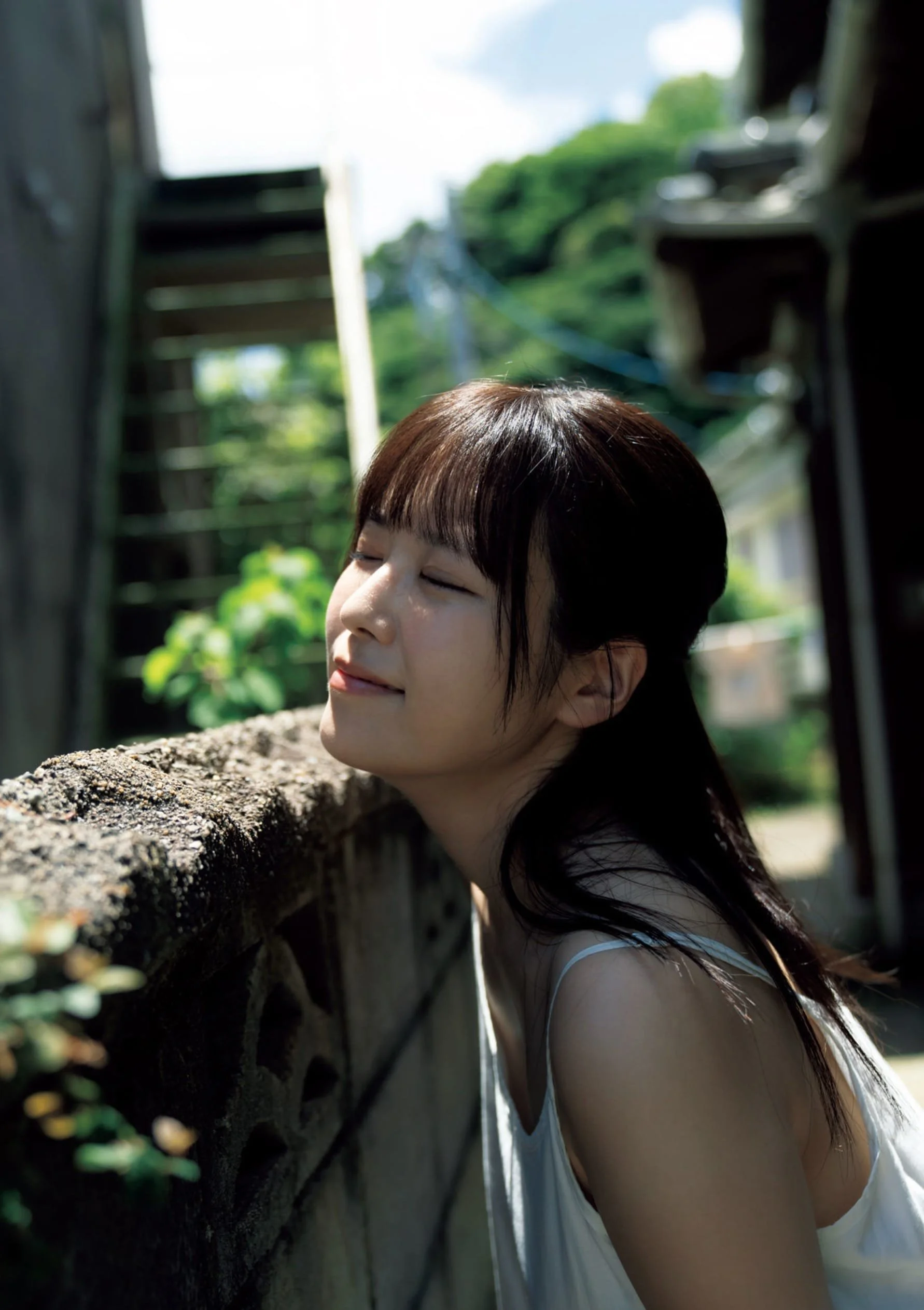 A woman with short dark hair and bangs is leaning her head against a textured stone wall outdoors with her eyes closed and a slight smile, illuminated by sunlight, with greenery and wooden structures in the background.