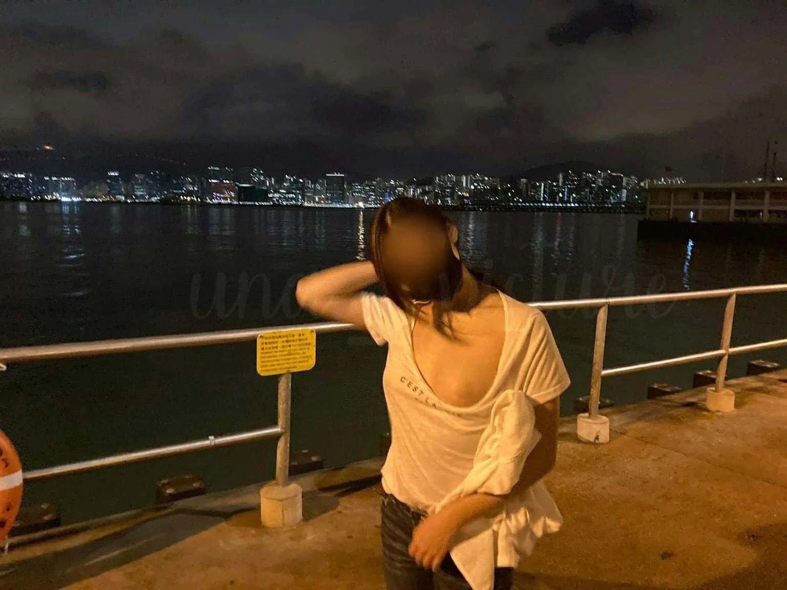 A woman stands by a waterfront railing at night with city lights reflecting on the water in the background. She is wearing a light-colored top and has her hand behind her head, with her face blurred.
