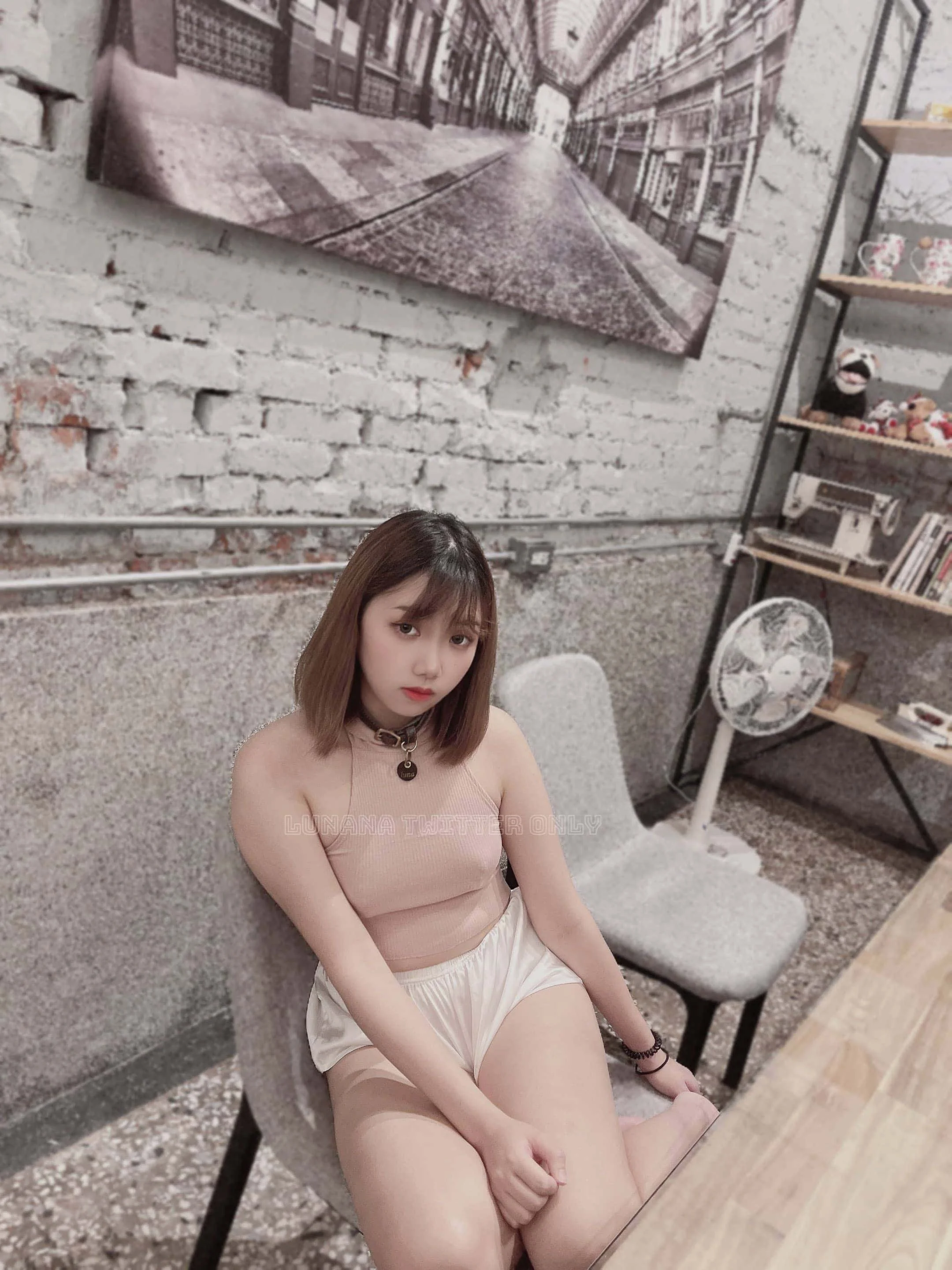 A young woman with shoulder-length brown hair sitting on a light gray chair in a room with exposed brick wall, a large black-and-white photograph hanging on the wall, a fan, and a bookshelf filled with various items.