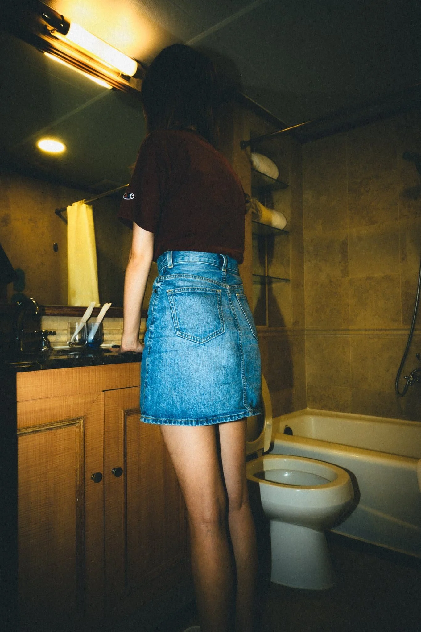 A woman wearing a maroon t-shirt and a denim skirt standing in a bathroom, facing away from the camera, with a bathtub and sink visible.