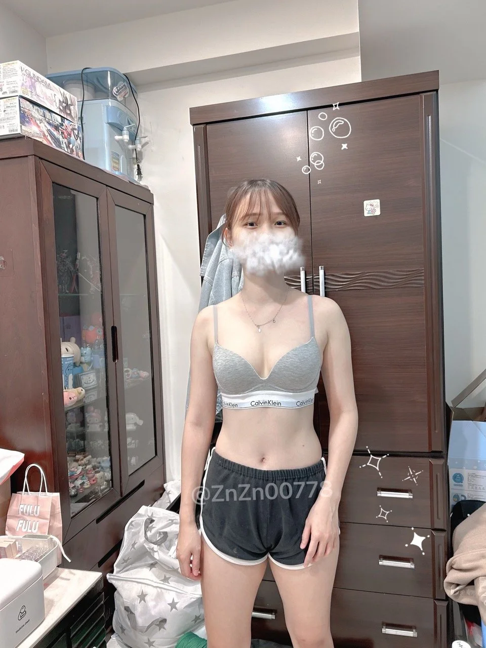 Young woman standing indoors, wearing gray Calvin Klein sports bra and black shorts, with digital sparkles and bubbles overlay, in a room with wooden furniture and various items.