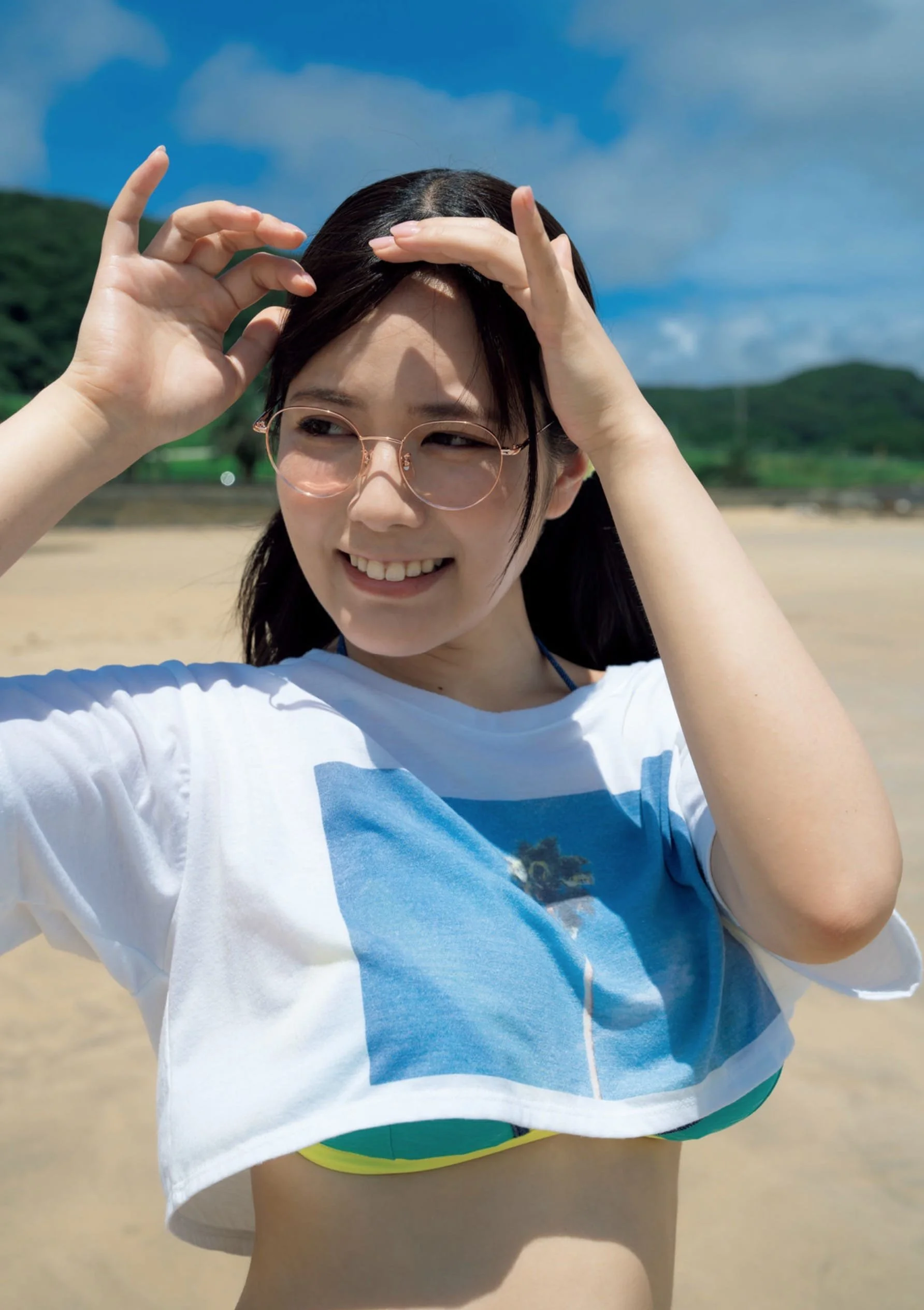 Young woman with glasses smiling and adjusting her hair outdoors on a beach with mountains in the background.
