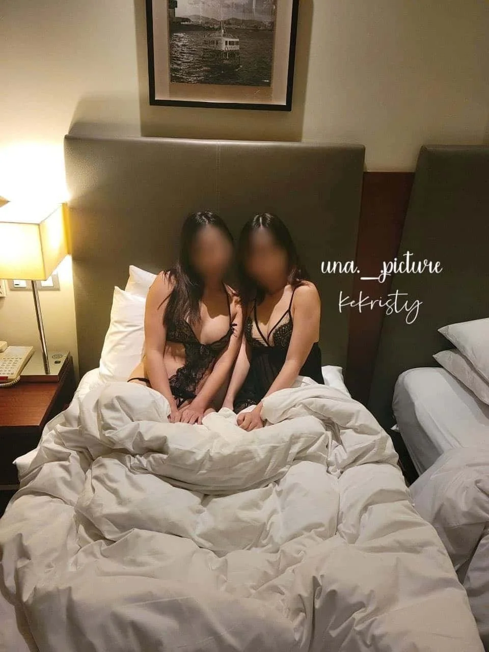 Two women dressed in black lace lingerie sitting on a bed with a beige headboard in a hotel room. An artwork hangs on the wall above them, and a nightstand with a lamp, phone, and clock is on the left side. The text 'una._picture' and 'kekristy' is o