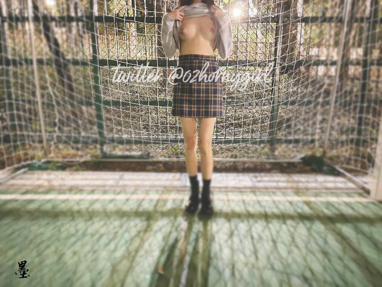 A person standing in front of a chain-link fence outdoors, lifting shirt to reveal bare chest, wearing a plaid skirt and black boots, with sunlight behind them.