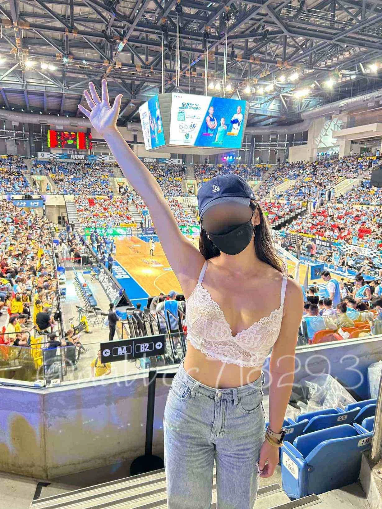 A woman wearing a black face mask, sunglasses, a navy blue cap, a white lace crop top, and light blue jeans standing in a sports arena, waving with one arm raised.