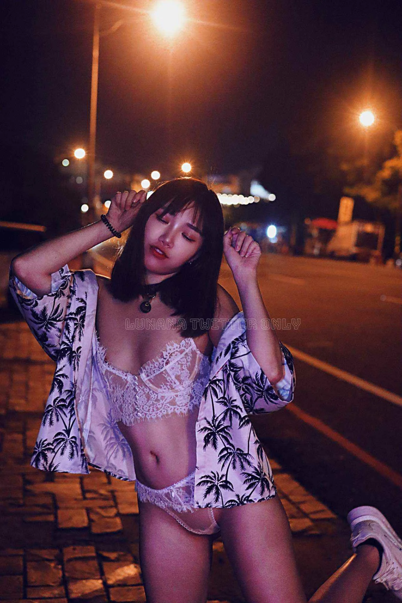 A young woman with black hair posing on a street at night, wearing a lace lingerie set, a patterned shirt, and sneakers, with streetlights illuminating the scene.