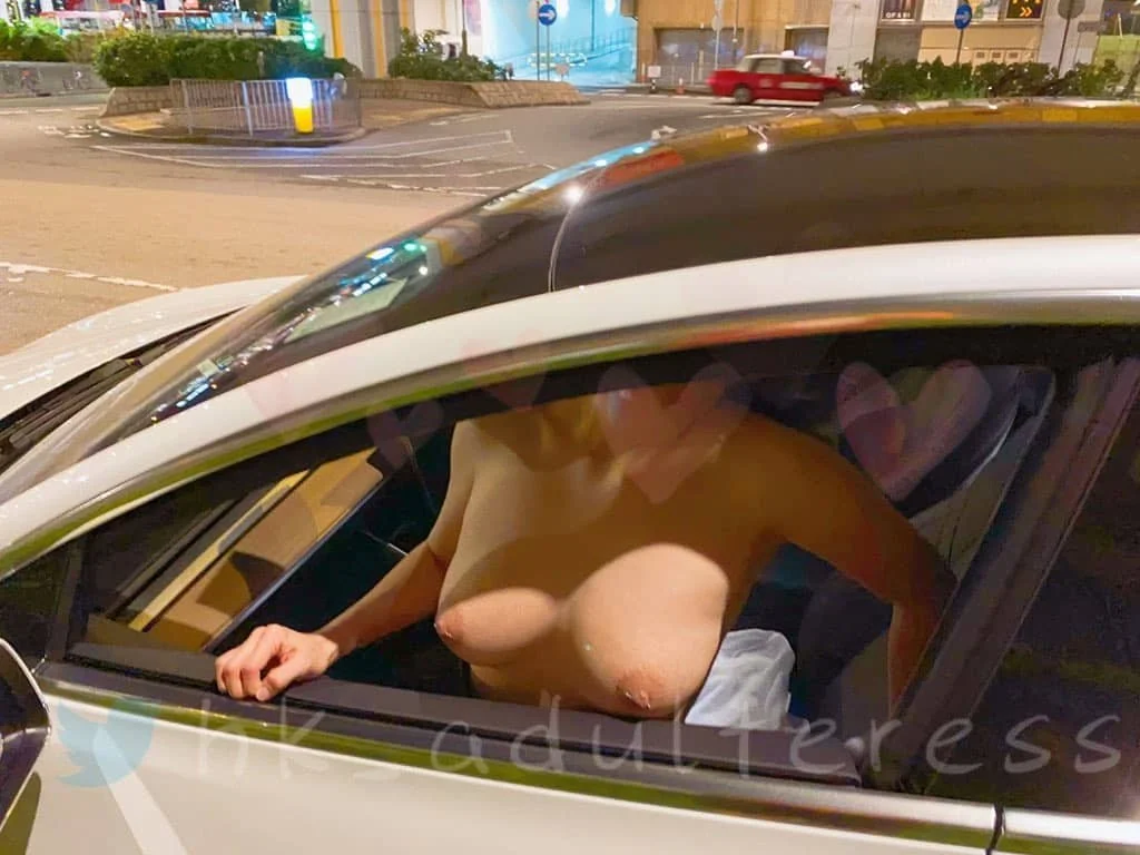 A topless woman sitting inside a car, viewed through the closed window. The urban street scene is visible outside the car.