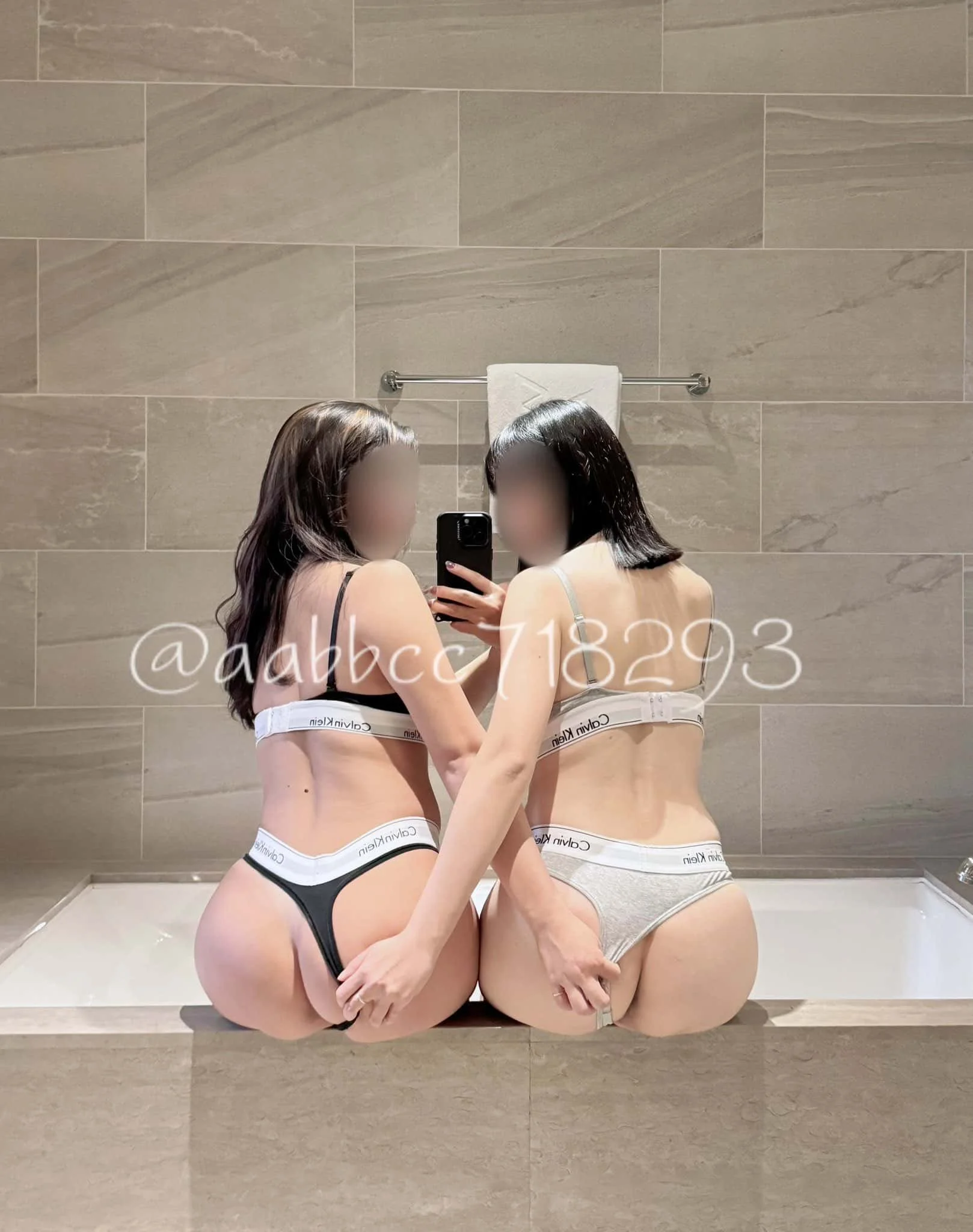 Two women in underwear taking a mirror selfie in a bathroom with beige tiles and towels.