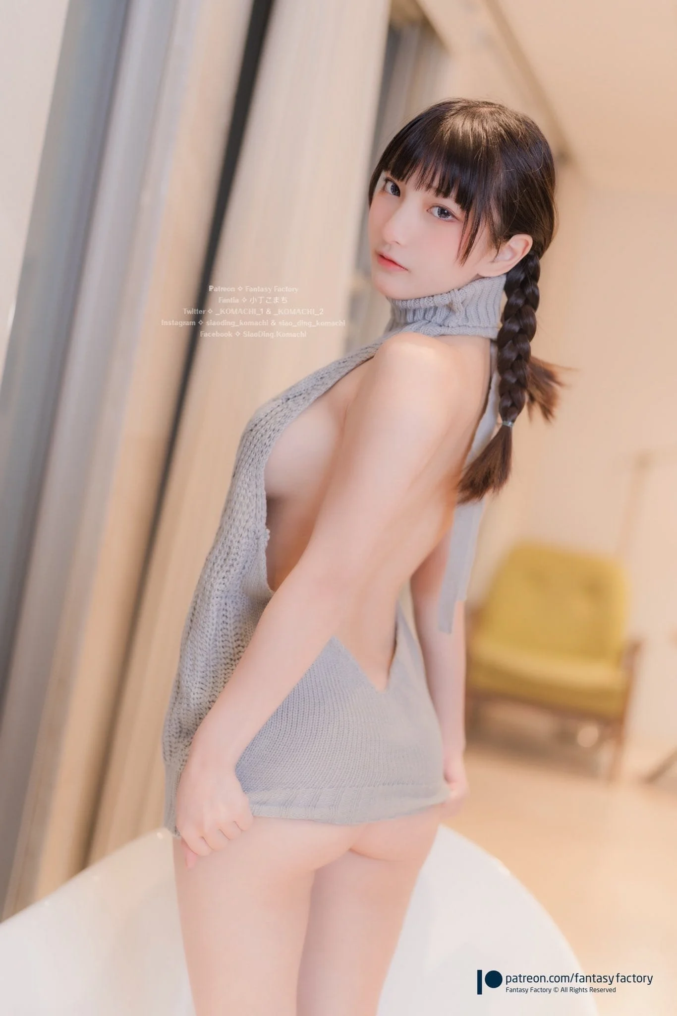 A young woman with short, dark hair styled in a braid, wearing a grey sleeveless turtleneck sweater that is partially pulled down to reveal her back, standing indoors near a wooden wall, looking at the camera.