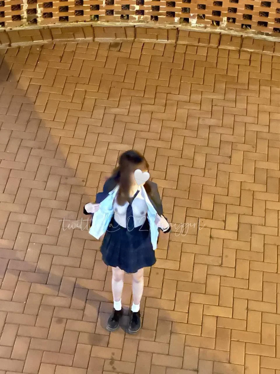 A girl wearing a school uniform standing on a brick sidewalk at night, with a brick wall in the background. Her face is obscured by a heart sticker.