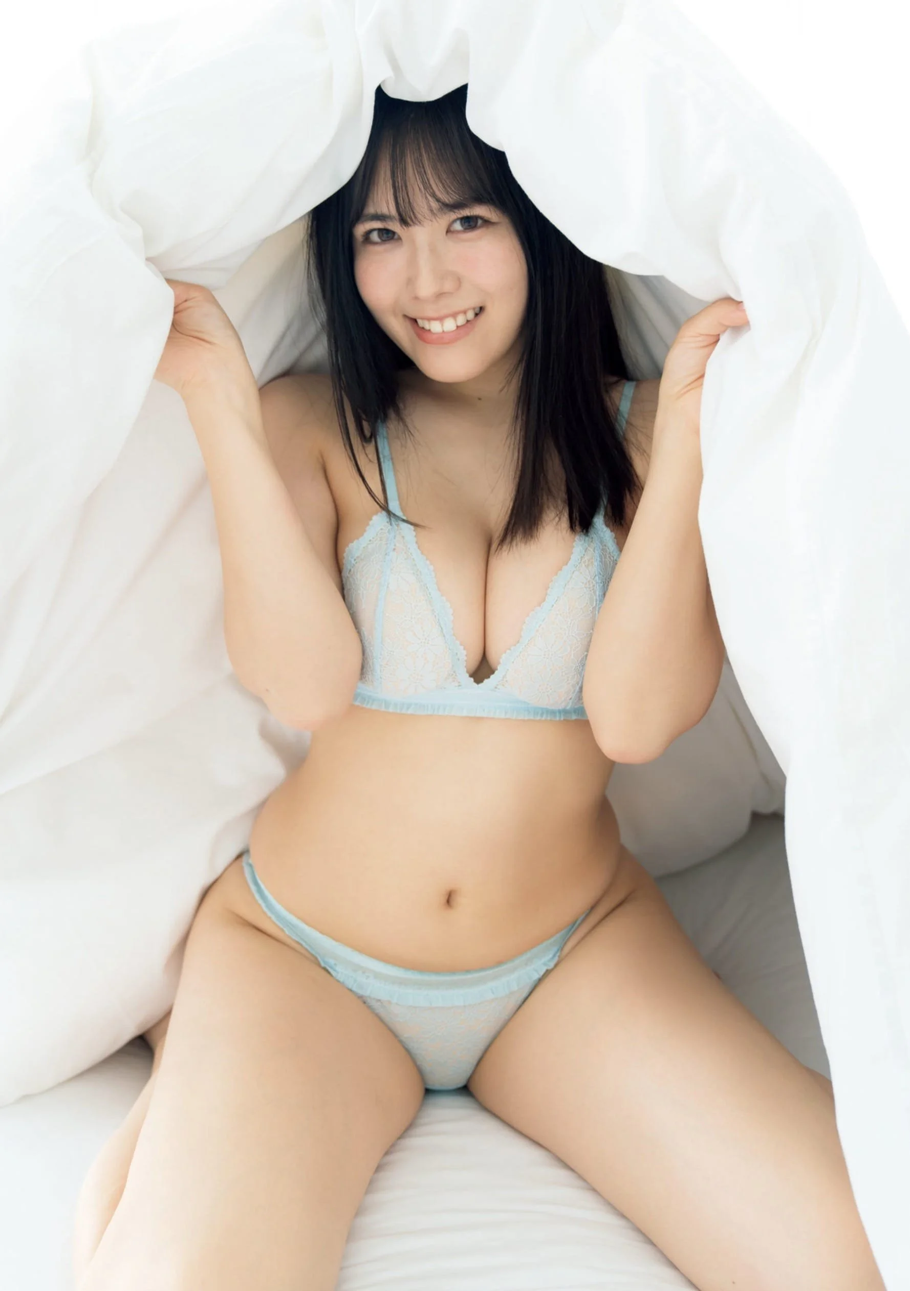 Young woman with dark hair smiling, wearing blue lace lingerie, sitting on a bed with white sheets and roofed by a white blanket.