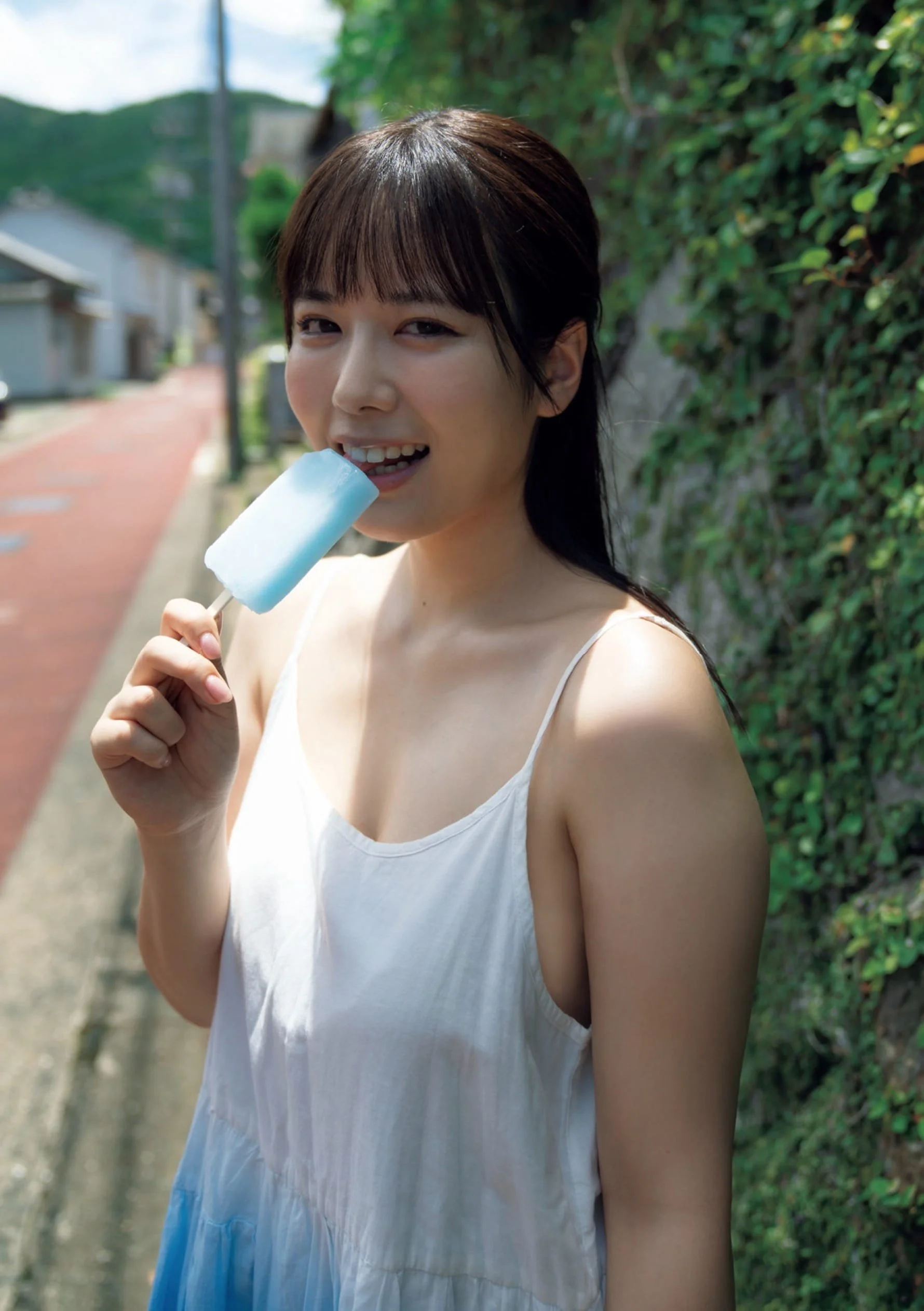 A young woman with dark hair and bangs wearing a white and blue sleeveless dress, standing outdoors on a sunny day, holding a light blue popsicle near her mouth, with a natural green hedge and a residential street in the background.