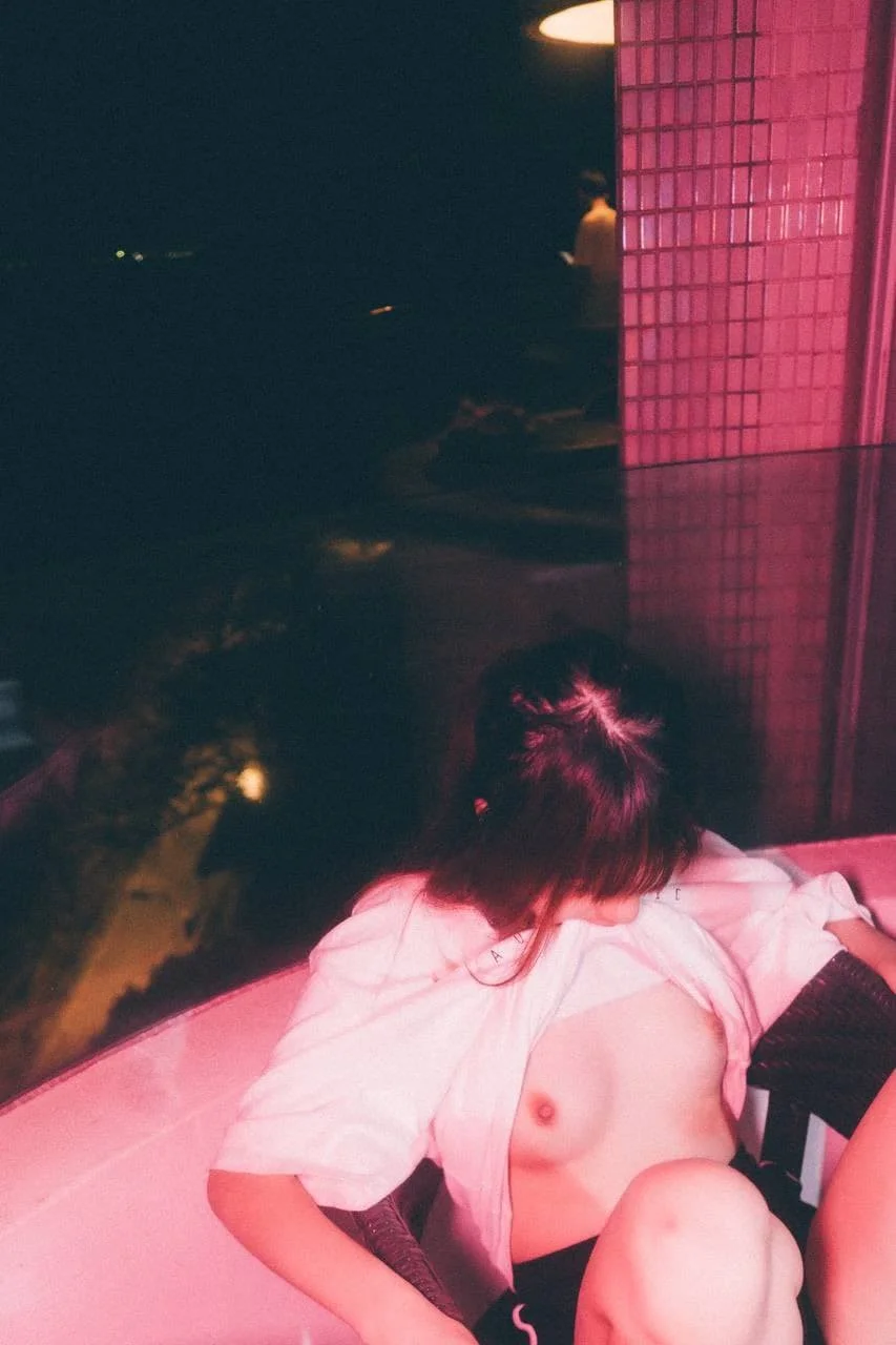 A woman with dark hair tied in a bun is sitting on a pink round couch with her shirt partially open, revealing her chest. The setting has pink and dark lighting, with a grid-patterned pink wall to the right and a dark background with lights visible i