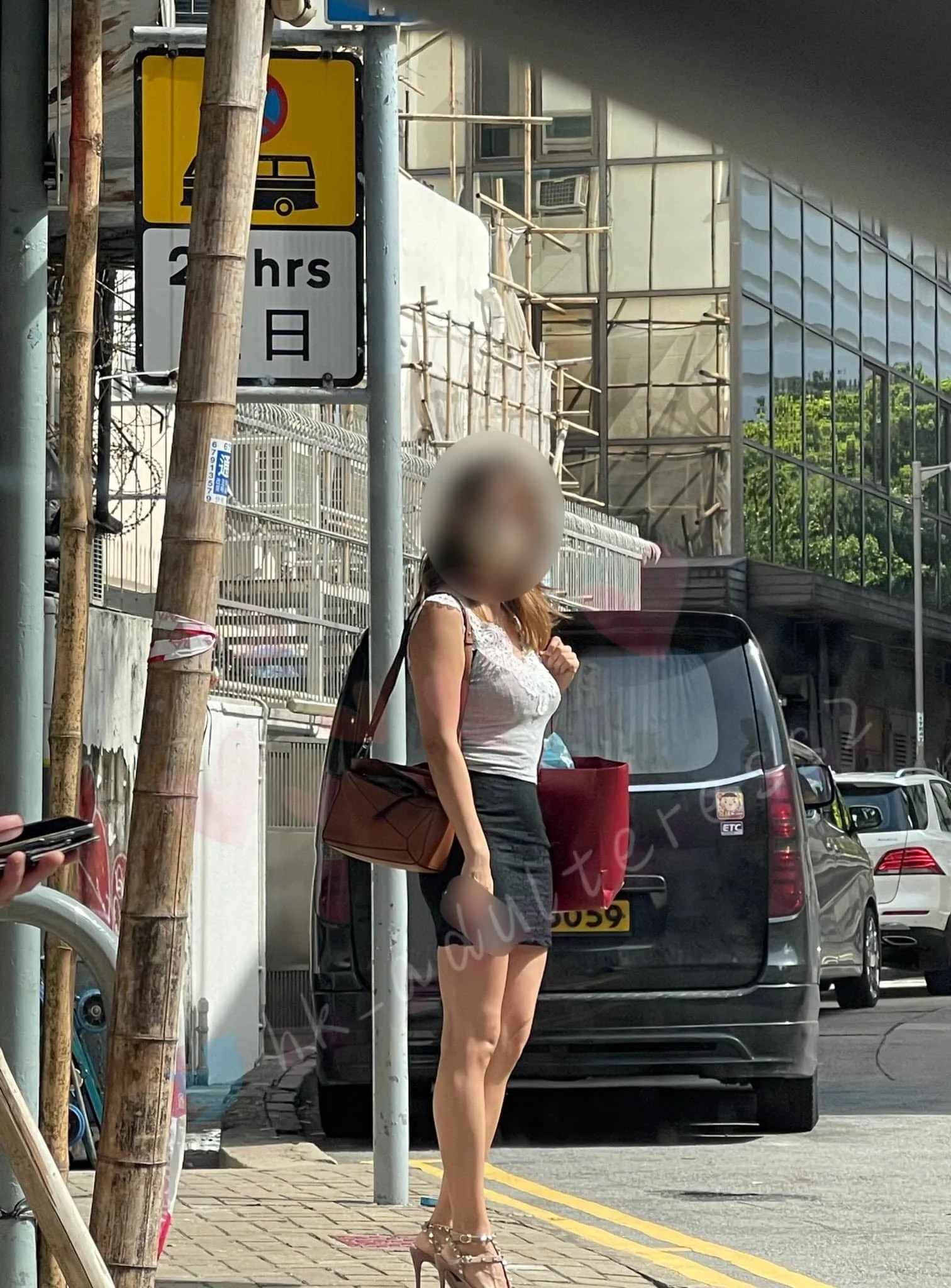 A woman standing on a sidewalk, carrying a brown bag and a red shopping bag, wearing high heels, a sleeveless top, and a black skirt, near parked cars and a street sign, in an urban area.