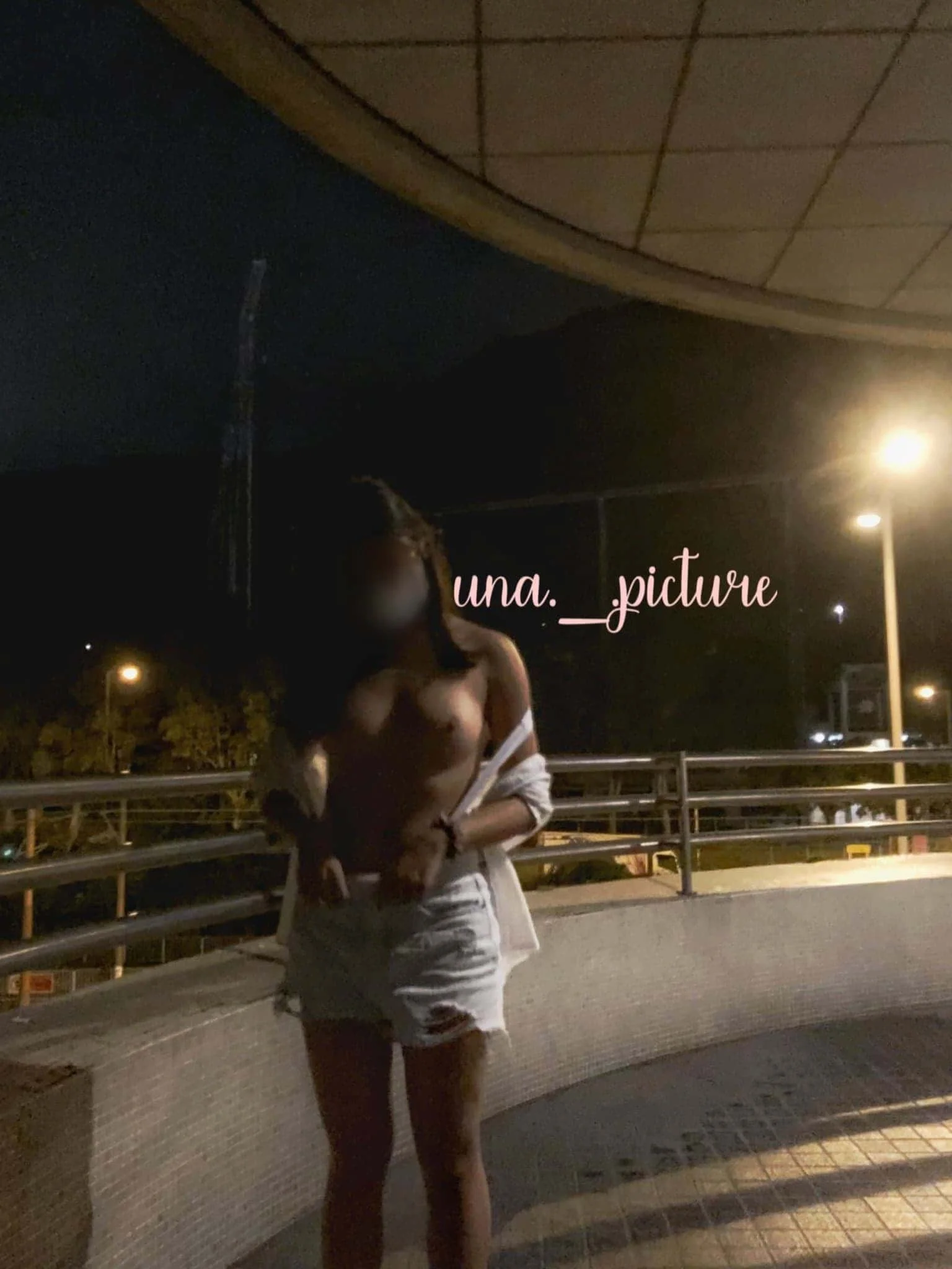 A woman stands on a dimly lit outdoor walkway at night, with streetlights in the background. She is topless, wearing shorts, and holding a jacket or shirt draped over her arm. The photo has pink text overlay that reads "una._picture".