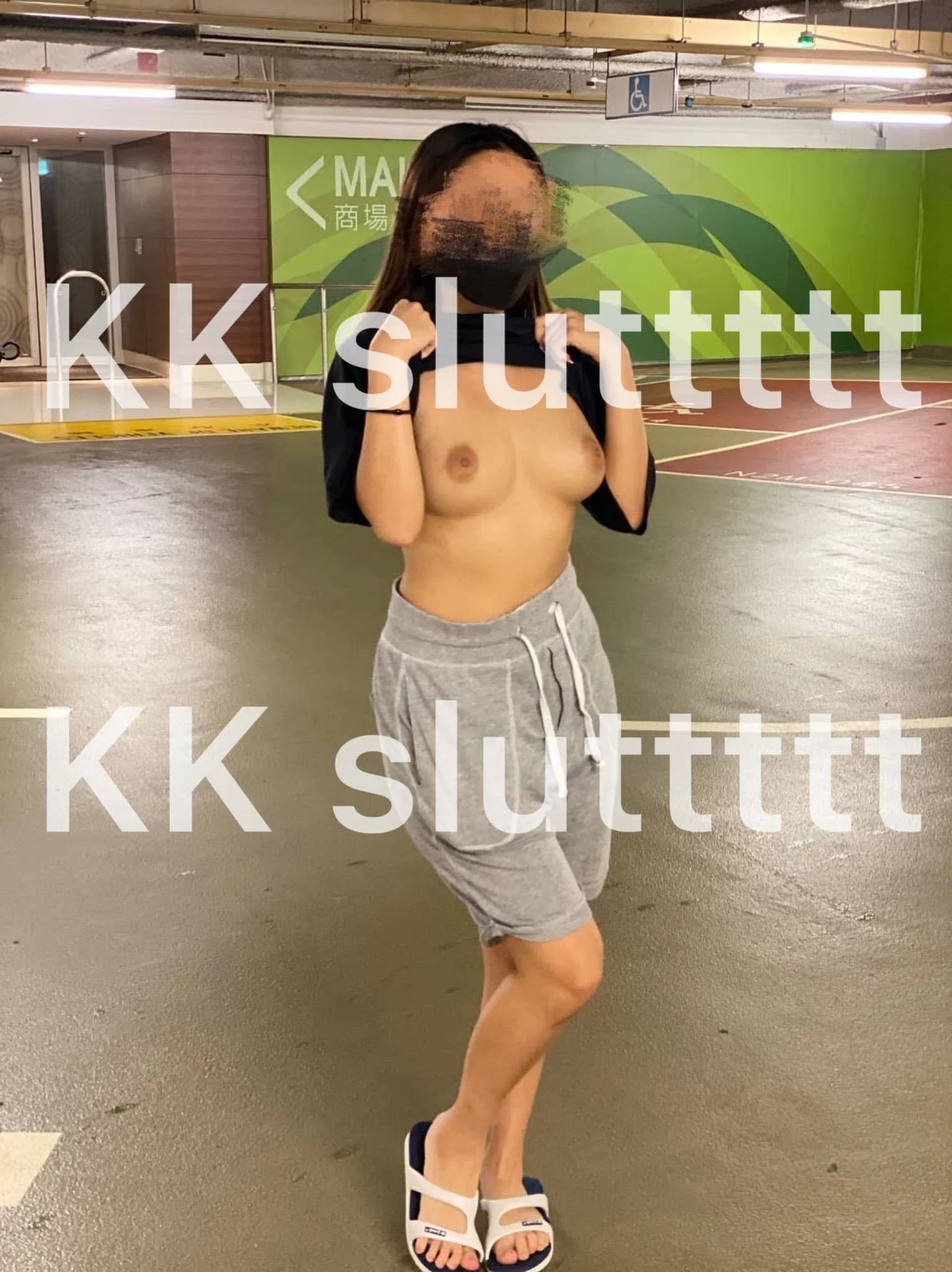 A woman in a parking garage lifts her shirt to reveal her bare chest, wearing gray shorts and white sandals.