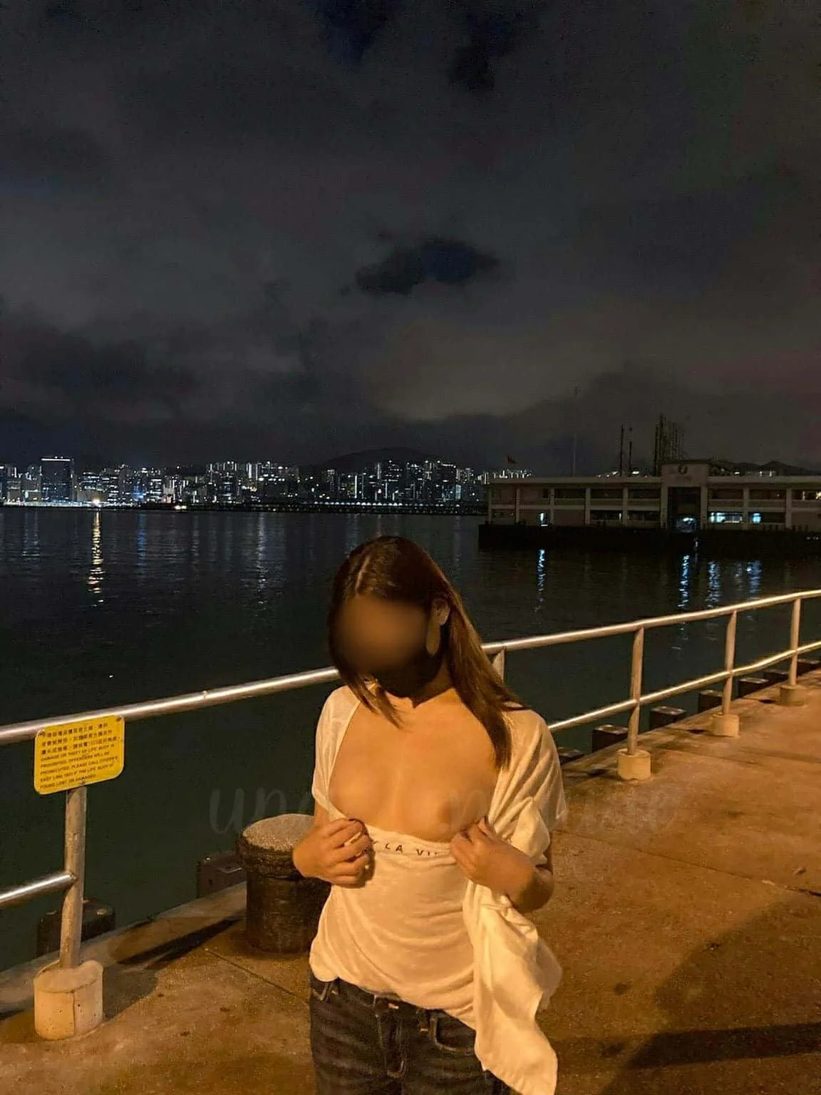 A woman standing on a pier at night, with city skyline and water in the background.