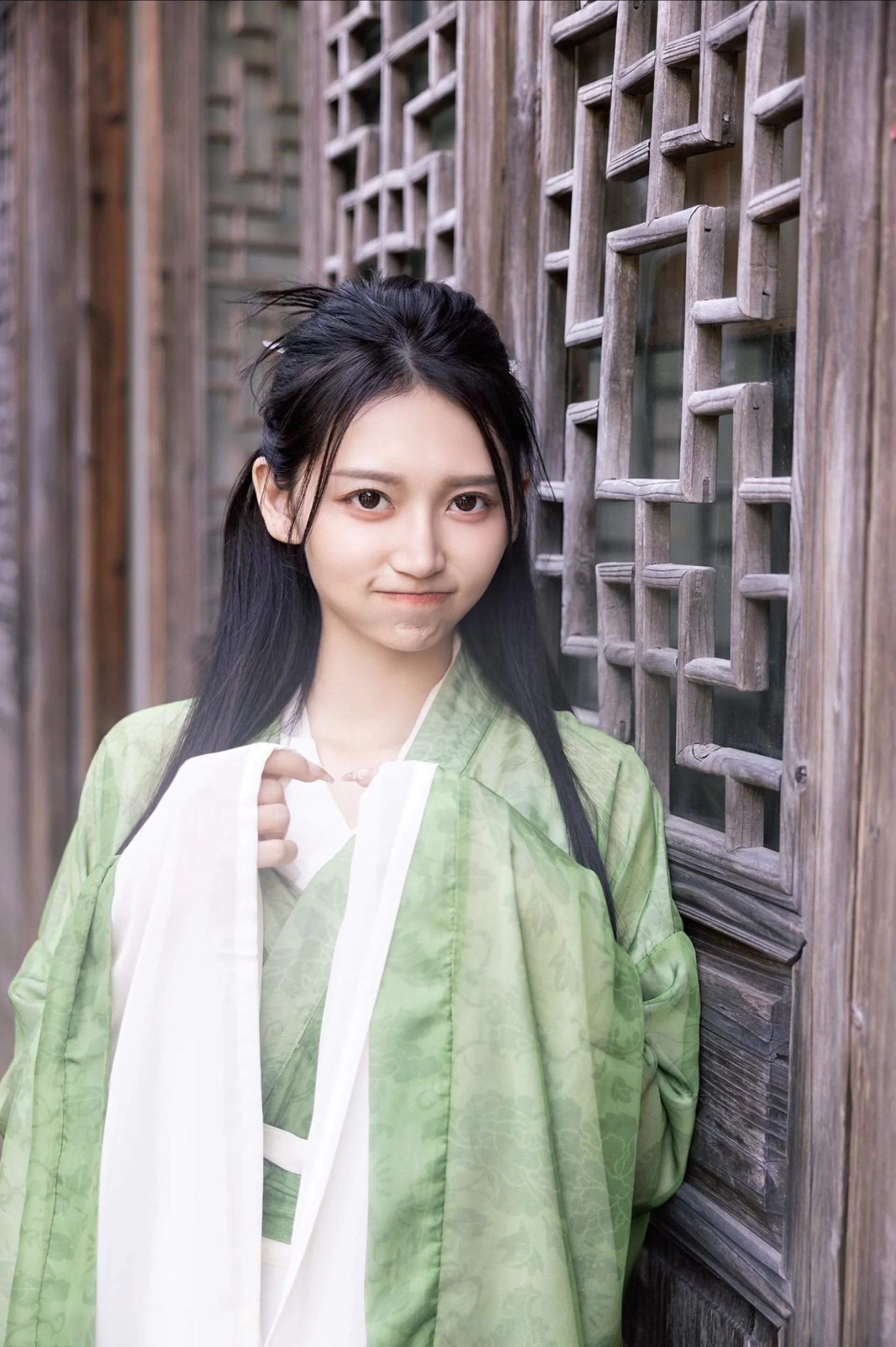 A young woman with long black hair, wearing a green traditional garment, standing next to a wooden lattice window, making a playful expression with her lip slightly pursed.