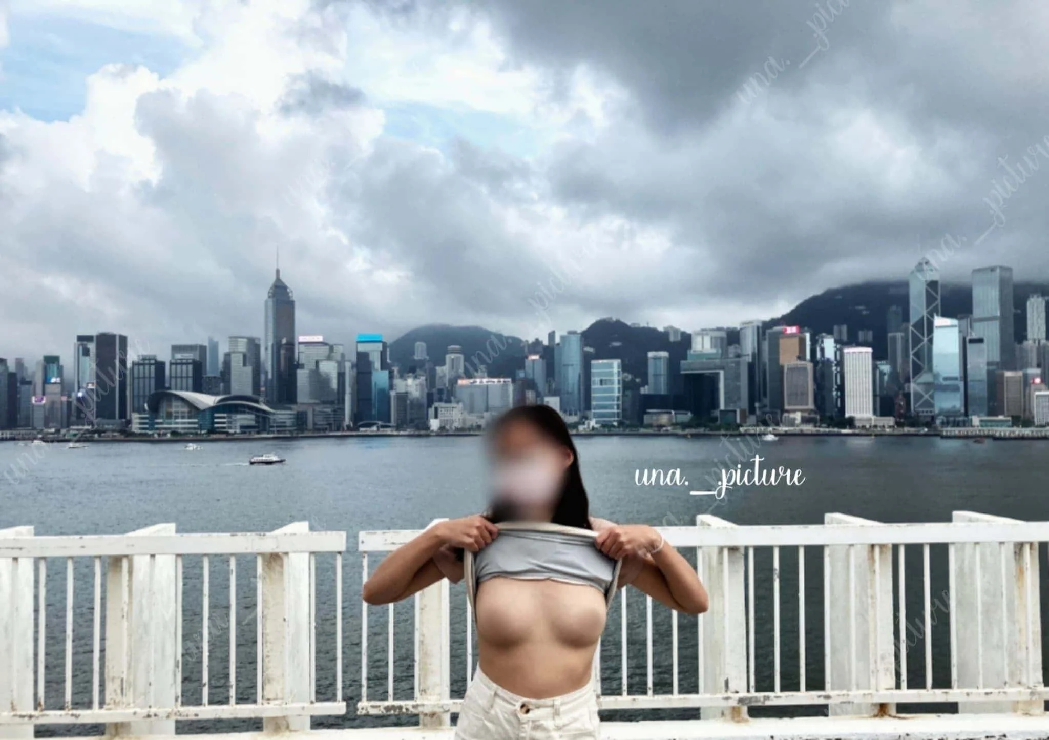 A woman with blurred face lifting her shirt on a waterfront balcony, with a city skyline and cloudy sky in the background.