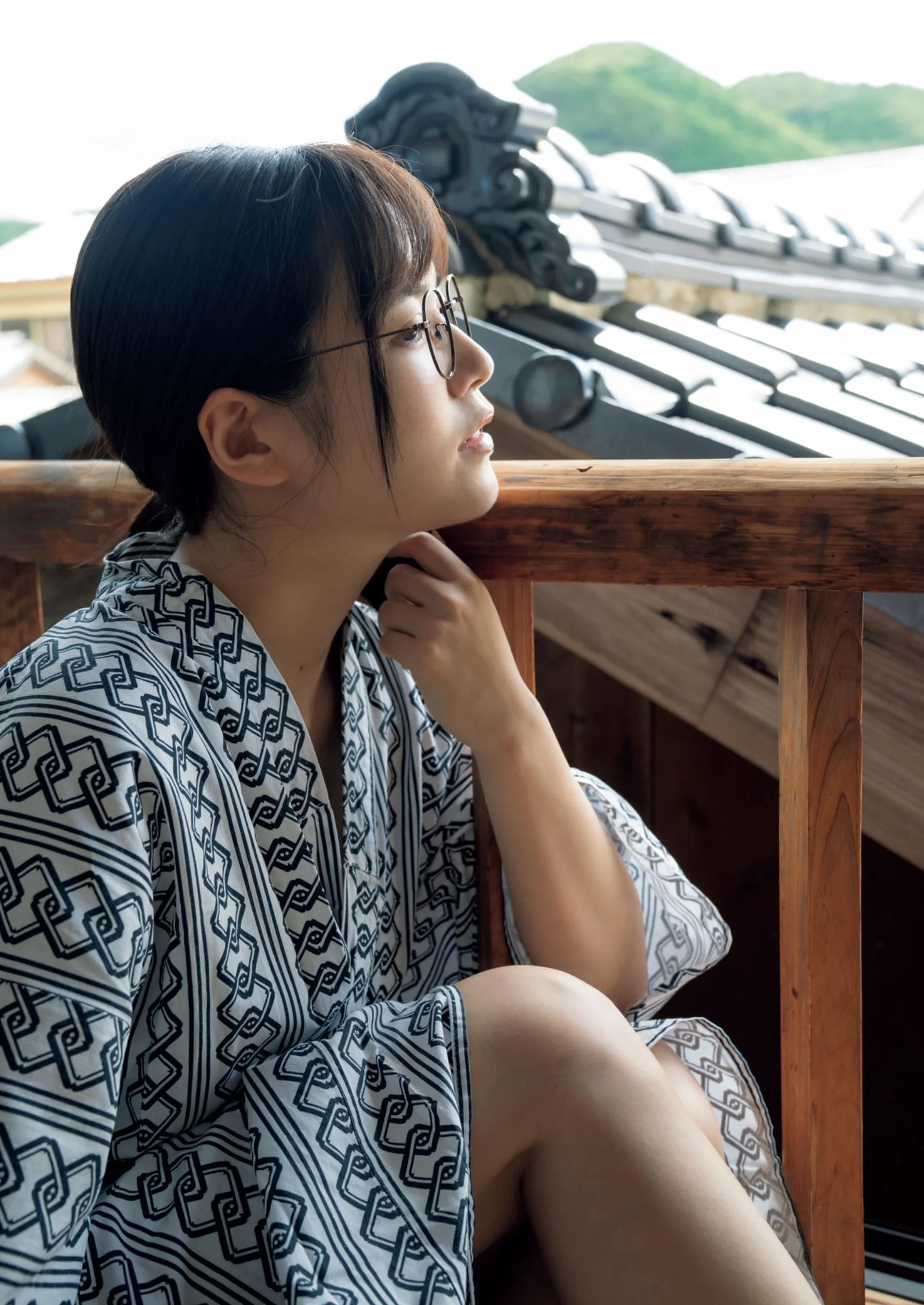 A woman with short dark hair and glasses sitting on a wooden porch, looking contemplatively into the distance with a traditional Japanese roof and green mountains in the background.