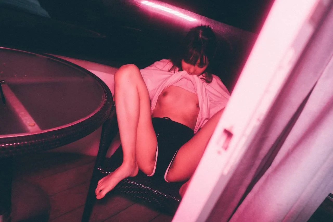 A woman is lying down on a chair with a pink light illuminating the scene. She is lifting her pink shirt to reveal her midriff and has her eyes closed.