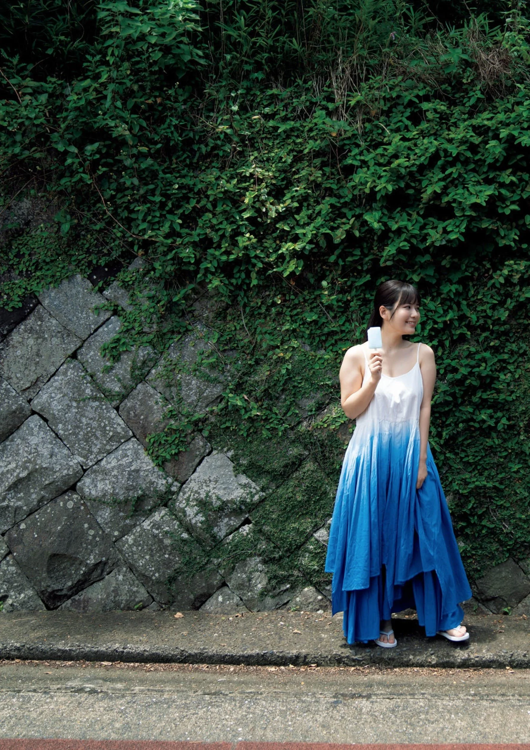 A woman in a flowing white and blue dress stands on a sidewalk holding a white popsicle, smiling and looking to her right, with a green leafy background and stone wall behind her.