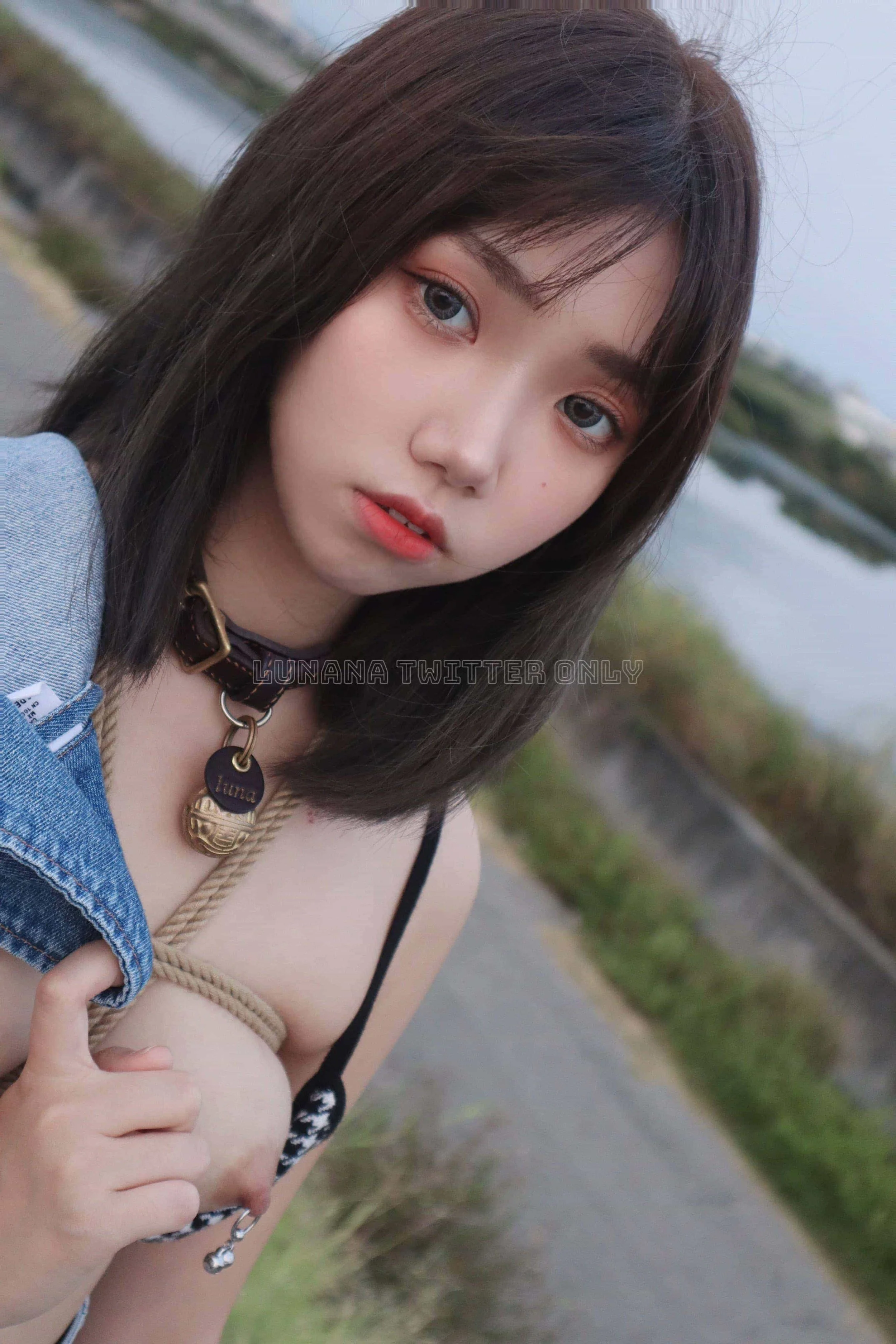 A young woman with shoulder-length dark brown hair and light eye makeup taking a selfie outdoors near a body of water. She is wearing a choker necklace with a pendant, a denim jacket partially off her shoulder, and a black and white top. The backgrou
