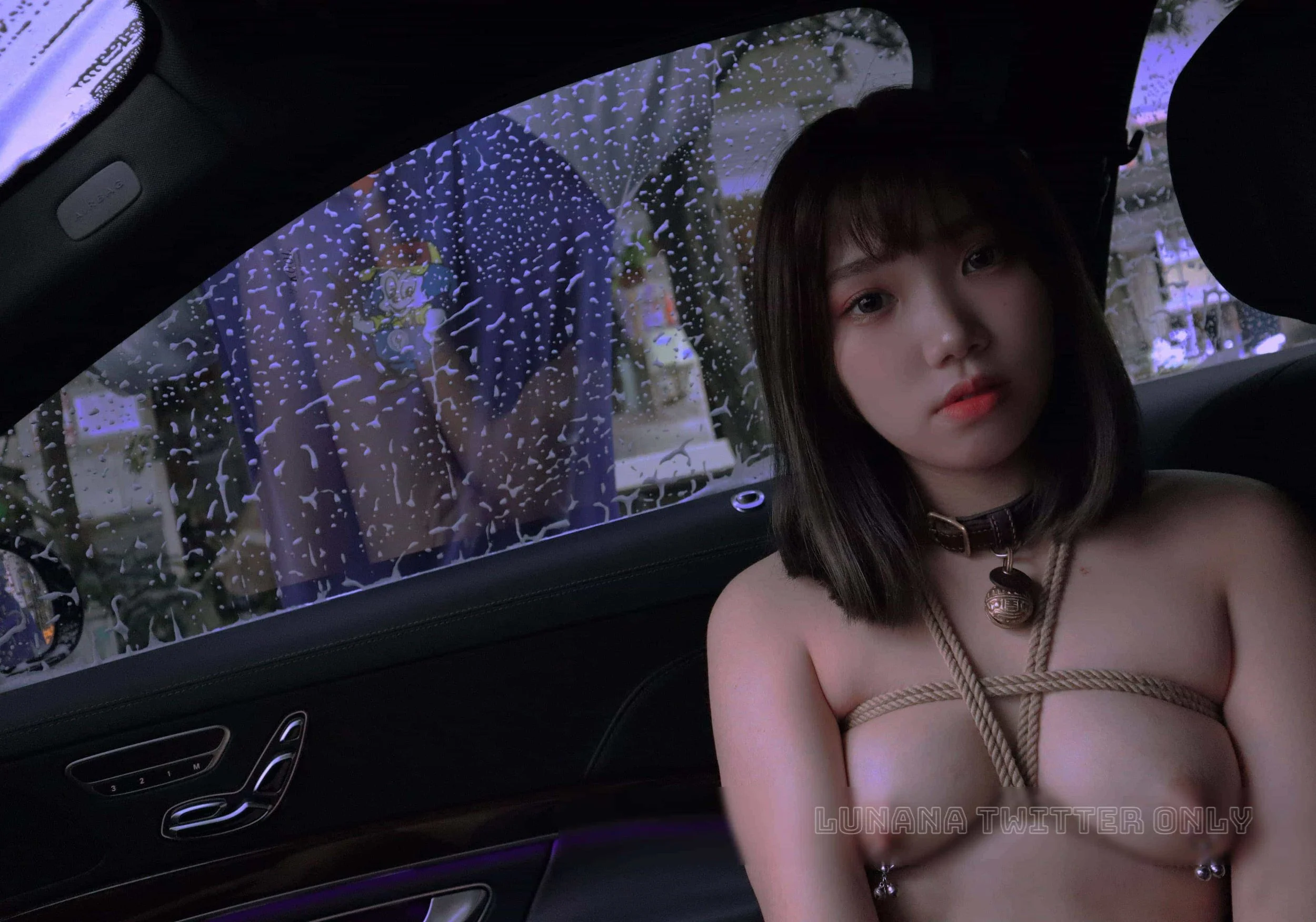 Young woman with shoulder-length dark hair, minimal clothing, and jewelry sitting inside a car, with rain on the window behind her and a person washing the car in the background.