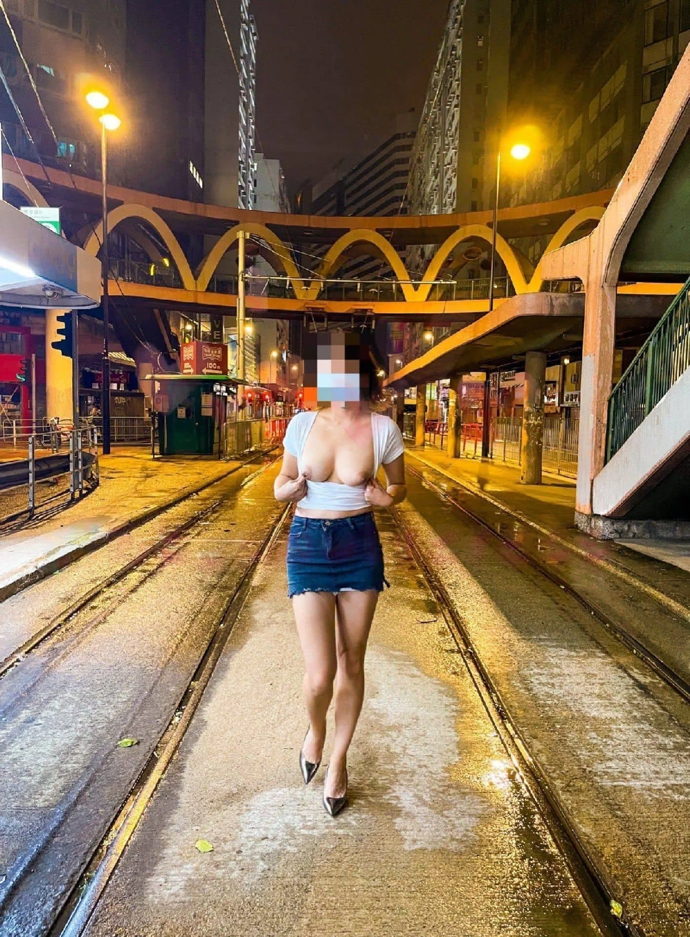 A woman with a pixilated face wearing a white crop top, denim skirt, and high heels walking on wet tram tracks at night in an urban city, illuminated by streetlights and surrounded by buildings and pedestrian bridges.
