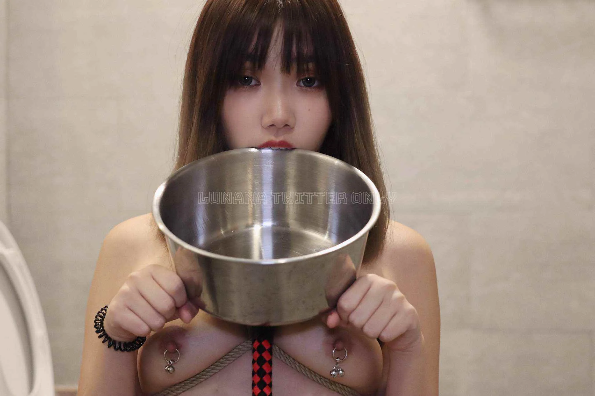 Young woman with dark hair and bangs holding an empty metal bowl close to her mouth, standing against a plain beige wall.