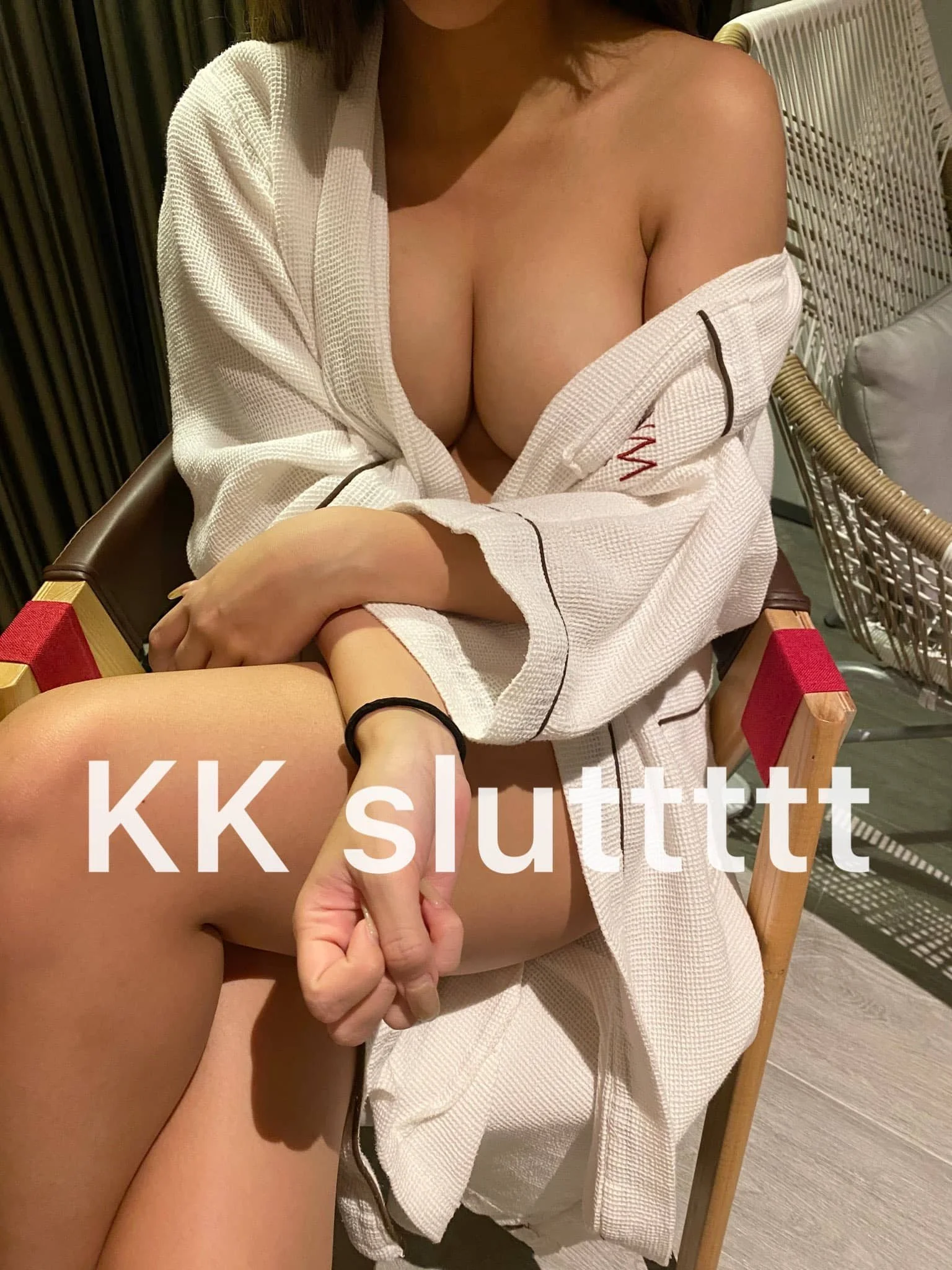A woman sitting on a chair with a white robe partly open, exposing her cleavage and legs, with the text 'KK sluttutt' overlaid.