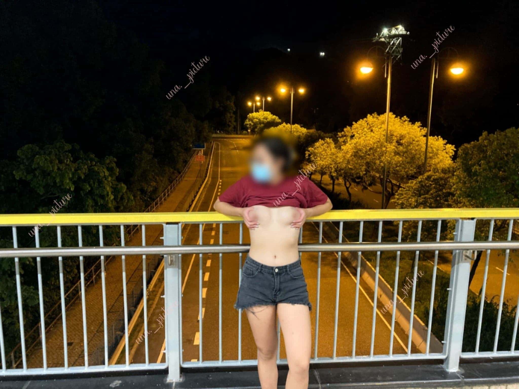 A person standing on a bridge at night with a blurred face, wearing a red shirt, black shorts, and a face mask, lifting their shirt to reveal their stomach, with a road and trees illuminated by streetlights in the background.