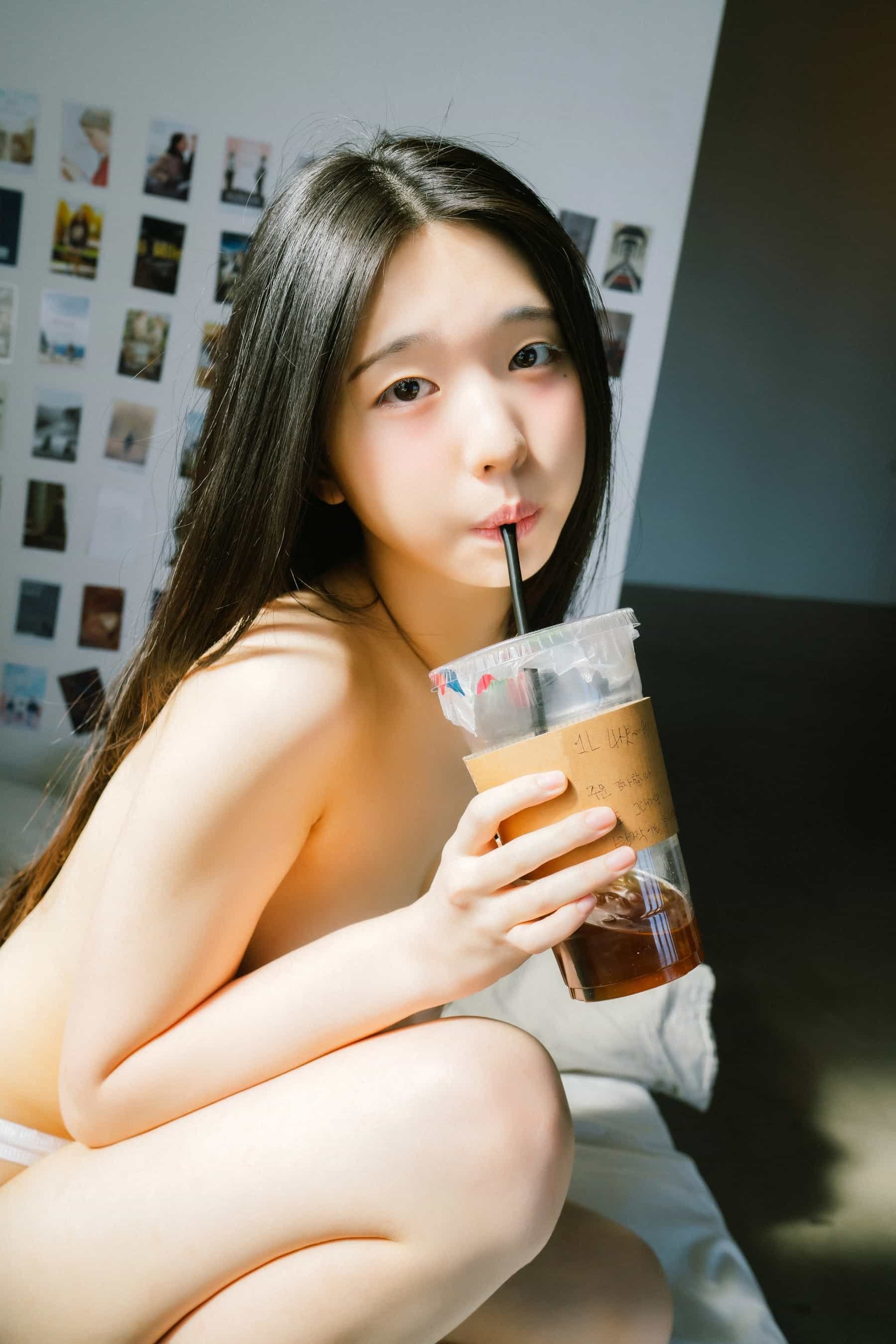 A young girl with long black hair sipping a cold drink through a straw, sitting on a bed with a wall of small photos or Polaroids in the background.