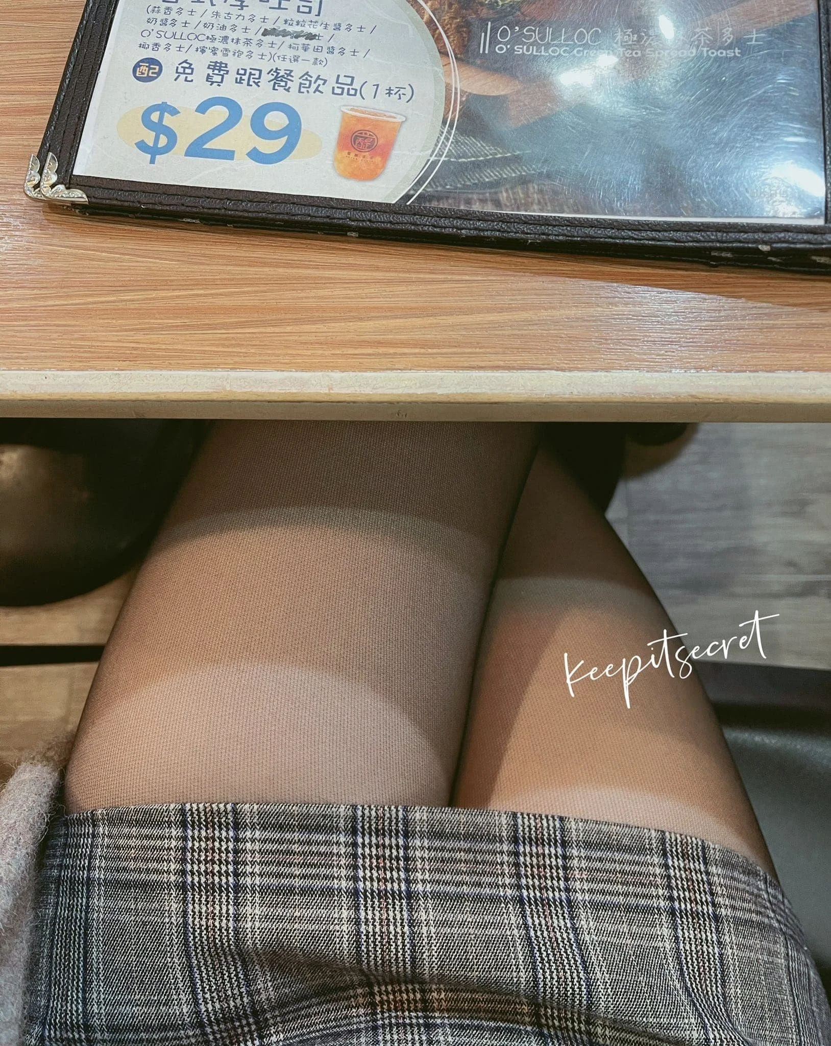 Close-up view of a woman's legs wearing beige sheer tights and a plaid skirt, sitting at a wooden table in a cafe, with a cafe menu placed in front of her.