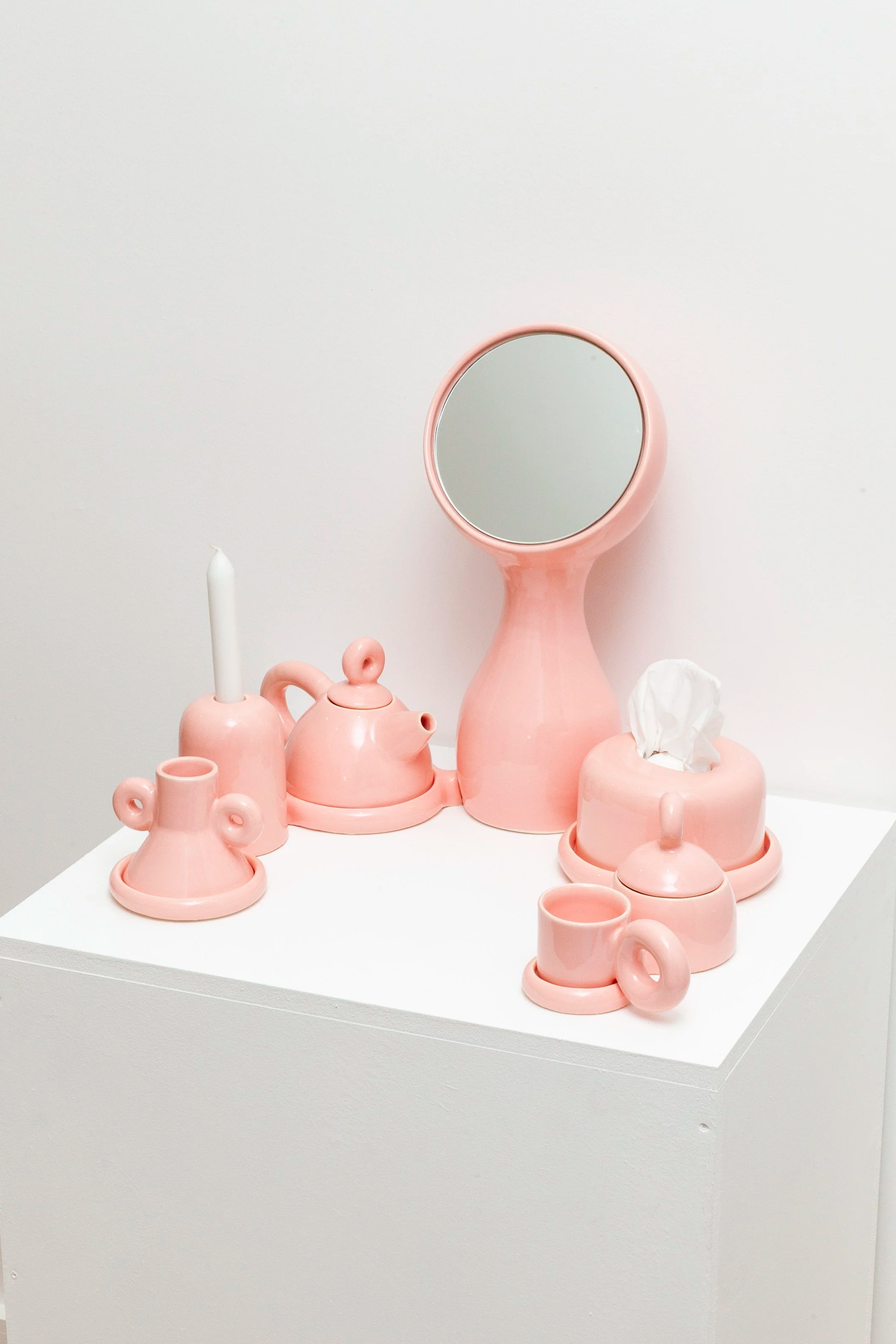 Pink ceramic accessories on a white pedestal, including a mirror, candle holder with a white candle, teapot, tissue box, and cups.