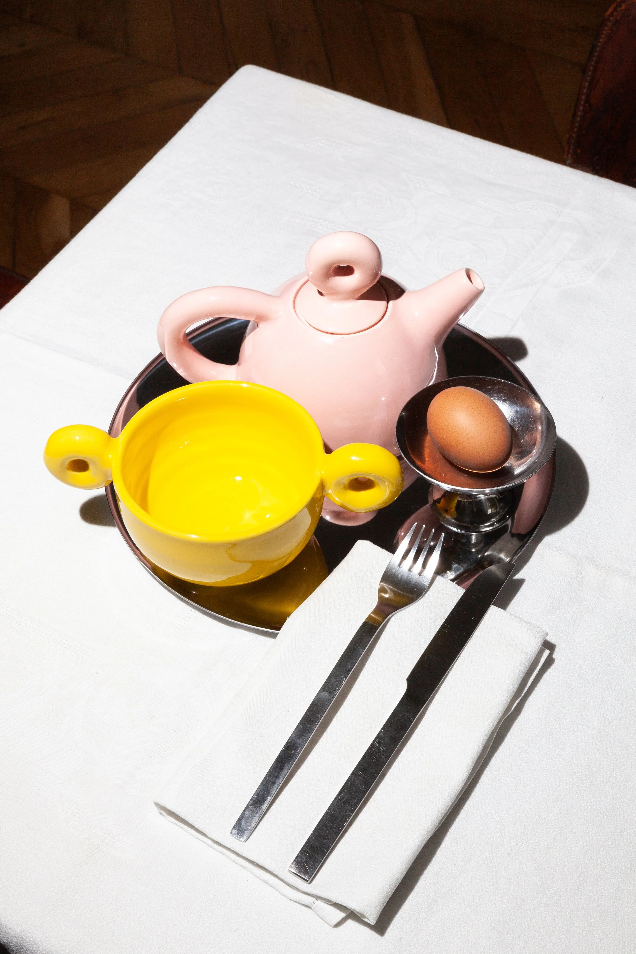 A table setting with a pink teapot, a yellow bowl, and a silver egg cup holding a brown egg. Silverware is placed on a white napkin on a white tablecloth.