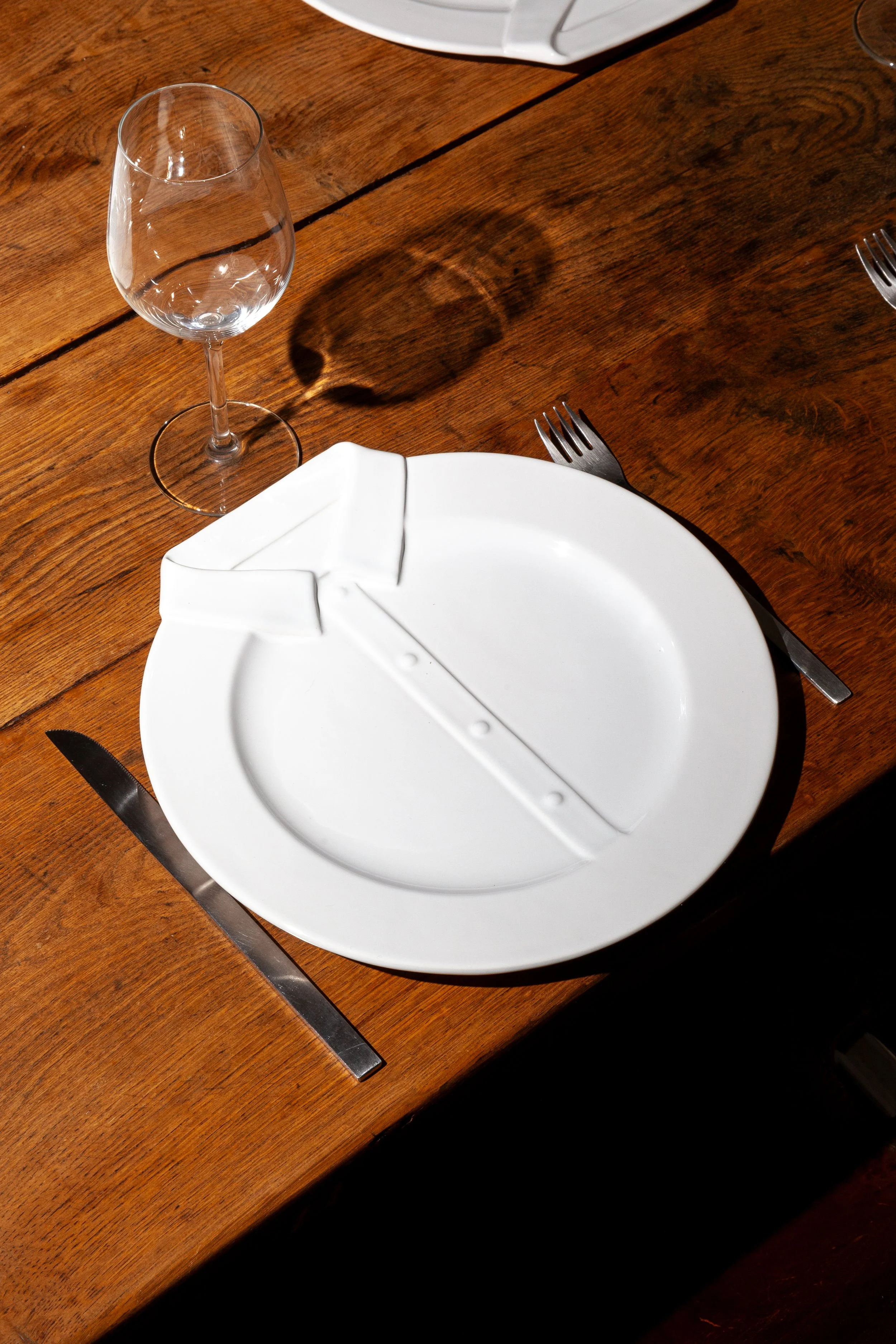 Empty white dinner plate with a built-in compartment for a salad or bread, set on a wooden table. There is a fork on the right and a knife on the left of the plate. An empty wine glass is placed above and to the left of the plate.
