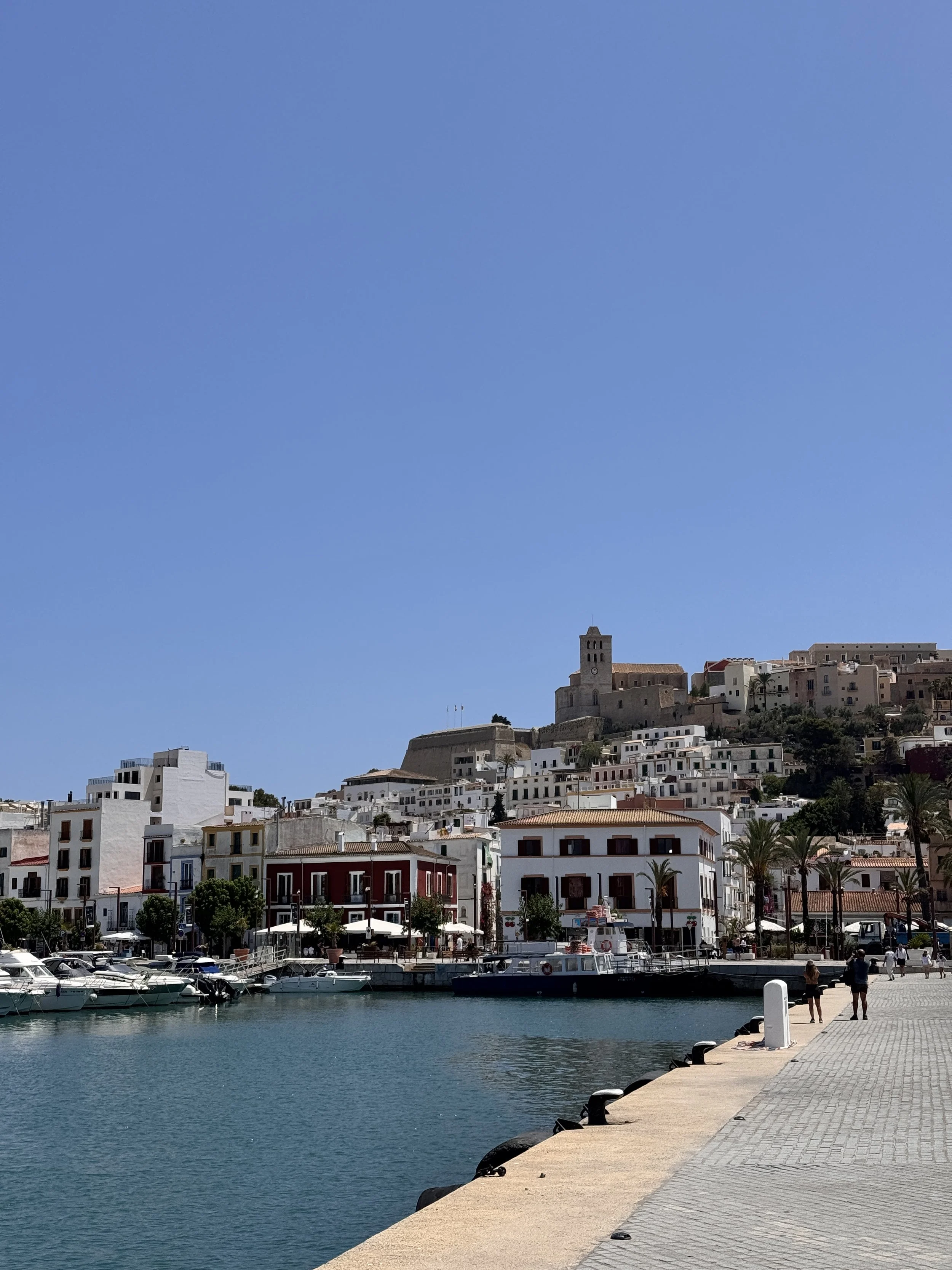 Ibiza old town. A scenic waterfront view of a small marina with boats, surrounded by white Mediterranean-style buildings, palm trees, and a hilltop historic tower under a clear blue sky.